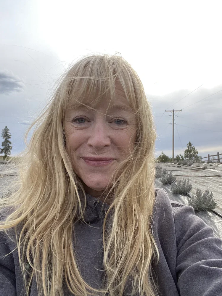A woman with long blonde hair taking a selfie outdoors on a cloudy day, with trees in the background.