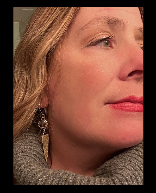 Close-up of a woman with blonde wavy hair, wearing a gray turtleneck sweater and dangling earrings, looking to the right.