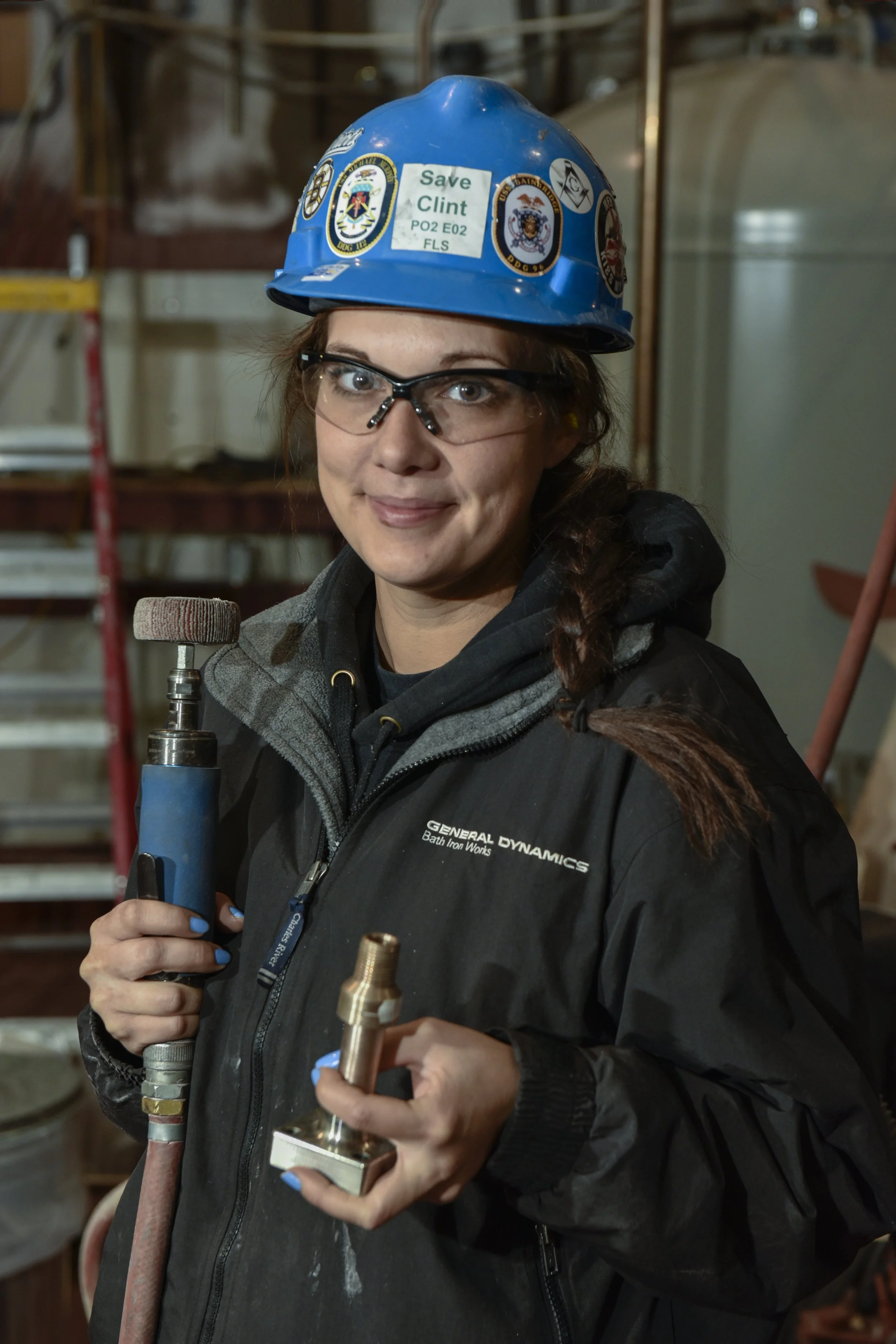 Machinist with hardhad and safety glasses poses for photo with a tool and metal part in her hands.