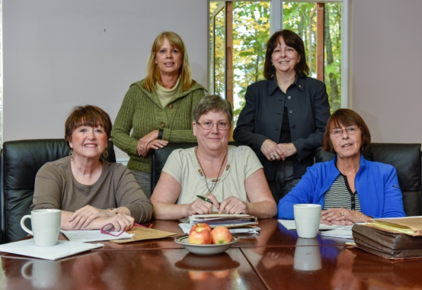 Five women gathered around a conference table in a room with large windows showing trees outside. 