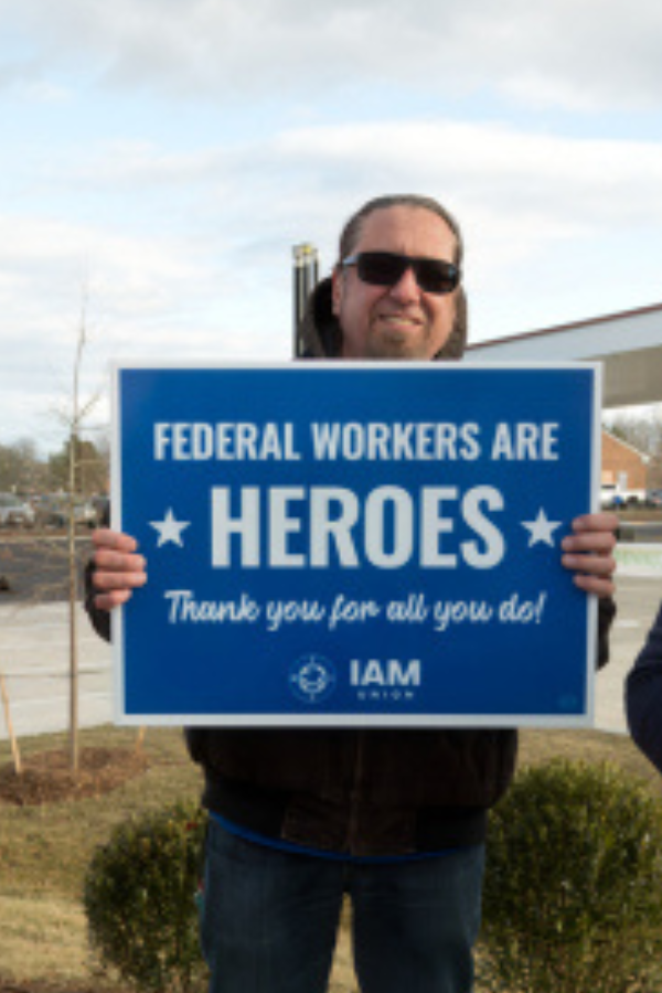 A man with a beard and mustache, wearing a light denim shirt, has his hand on his chin as if thinking, with a serious expression against a black background.