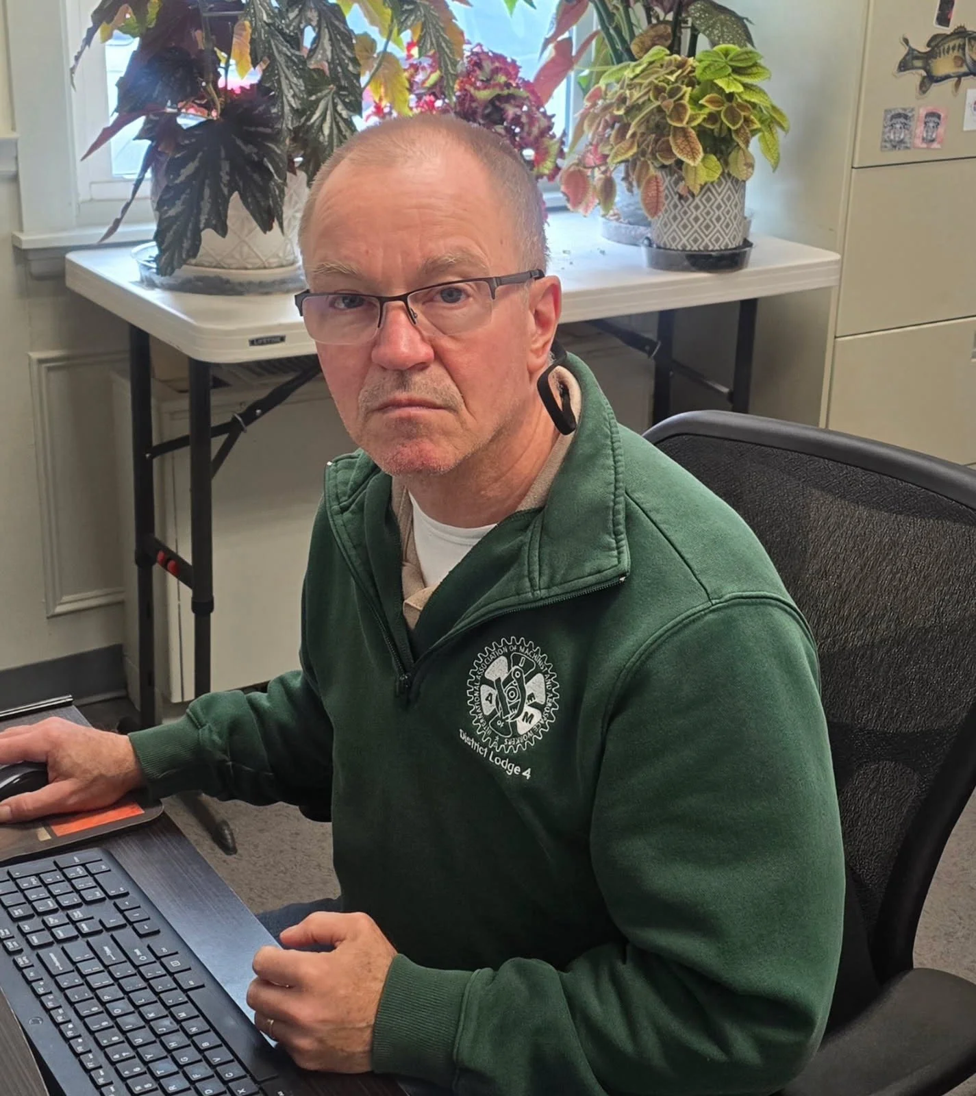A man with glasses, wearing a green jacket with a logo, sitting at a desk with a computer mouse and keyboard, in an office with potted plants in the background and a window.