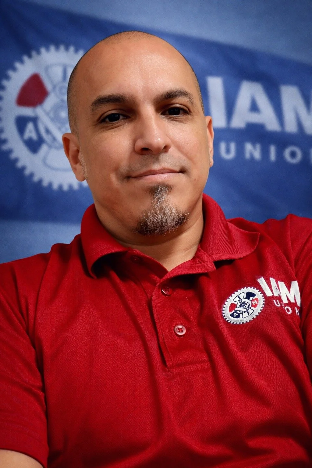 Portrait of a young man with brown hair, beard, and mustache, wearing a textured gray sweater, against a black background.