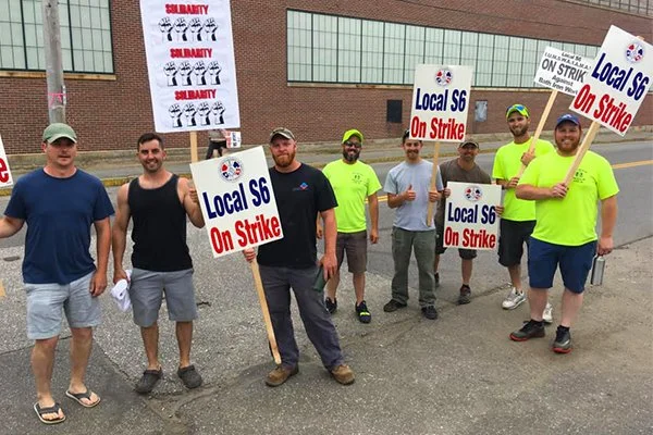 A group of seven protesters standing on a sidewalk holding signs that read "Local S6 On Strike." Some of the signs also have the word "SUBURBARY" and cartoon illustrations. The protesters are wearing casual clothing, with a few in bright yellow shirts, and are standing in front of a building with large windows.