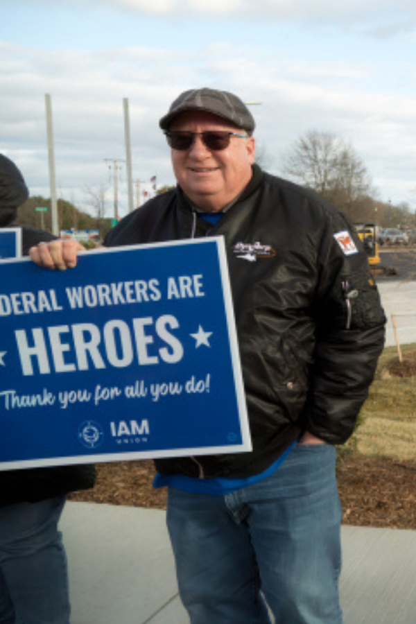 A smiling man with dark hair and a beard wearing a white shirt, standing against a blurred gray brick wall background.