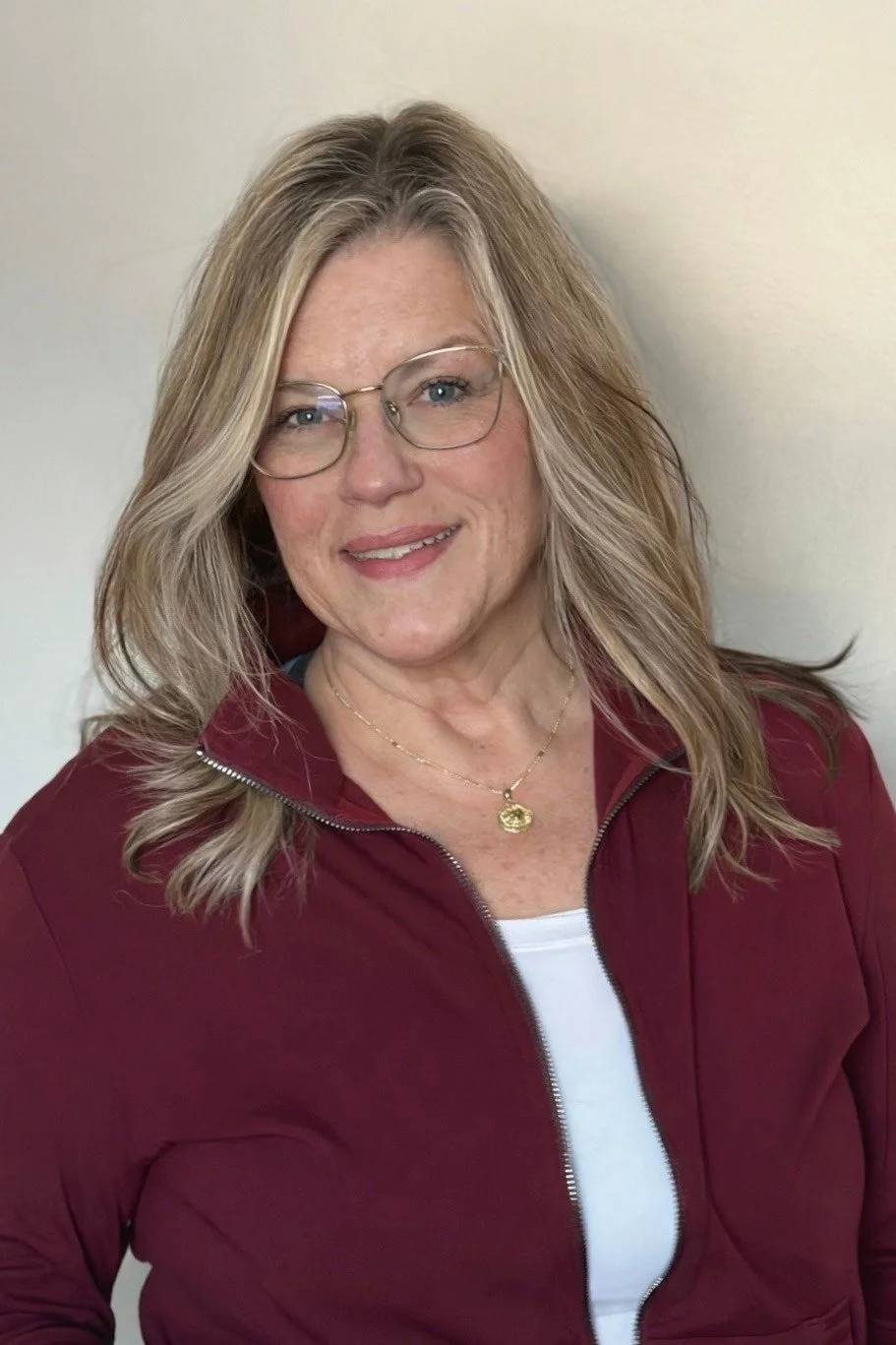 A woman with dark hair tied in a messy bun, smiling, with a nose ring, wearing a white shirt, against a plain white background.