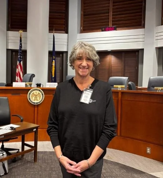 A woman with gray hair and a black top standing in a formal government or council chamber, smiling with an American flag and official seals in the background.