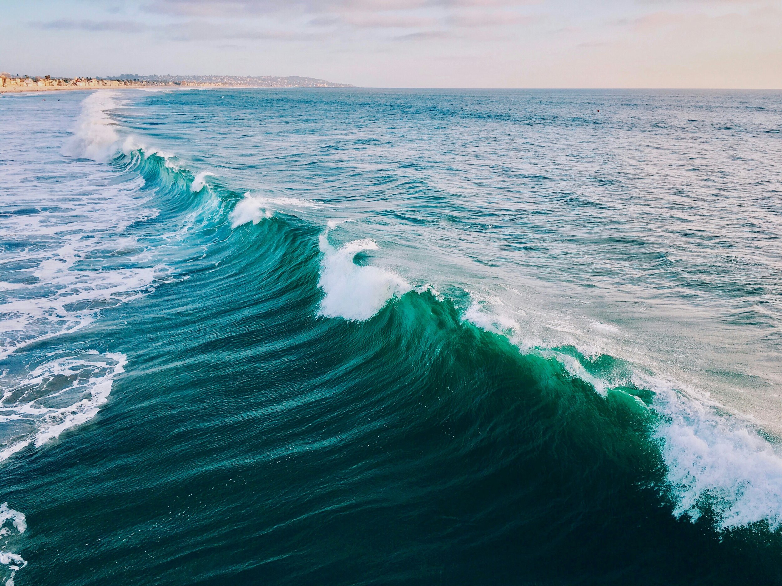 Ocean waves crashing near the shoreline with distant cityscape and clear sky.