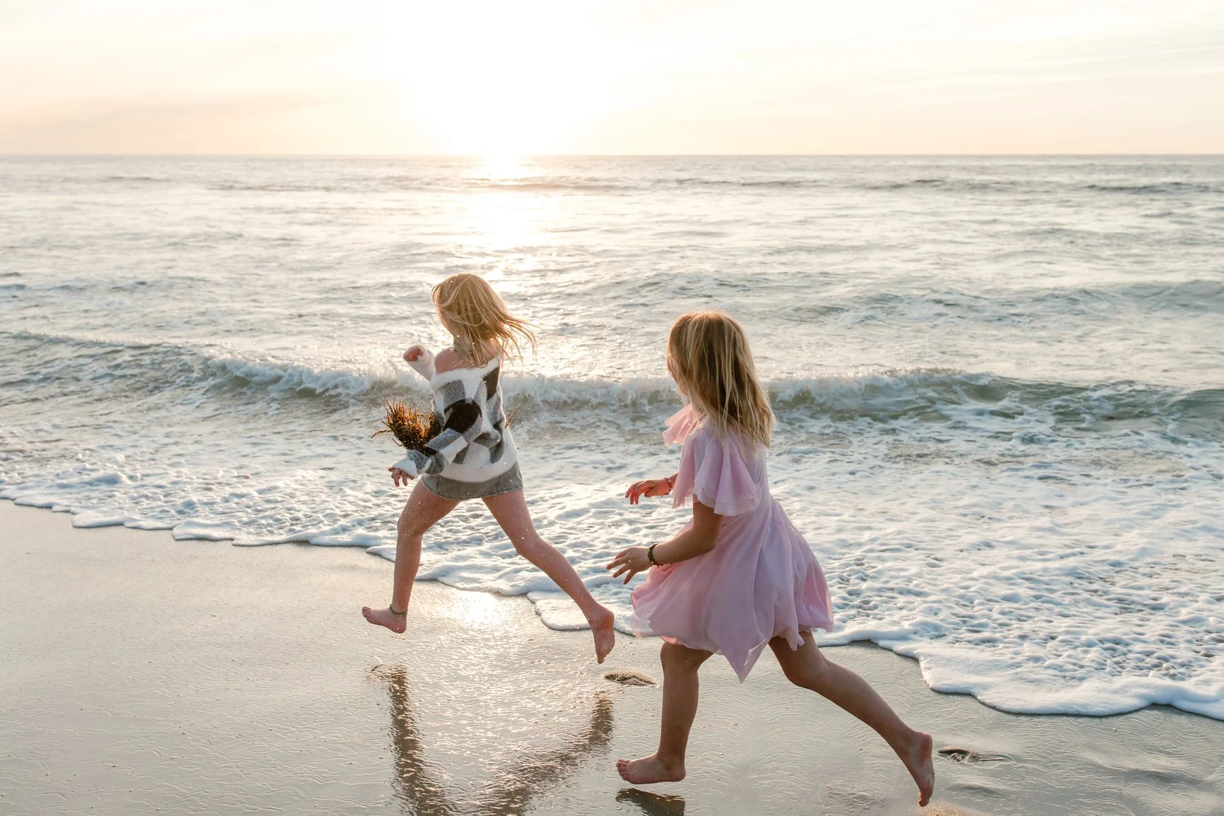 Two young girls playing and running on the beach near the water during sunset, with waves in the background.