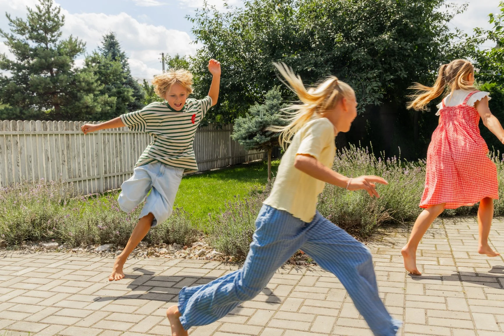 Three children are running playfully on a paved backyard with a white fence, green trees, and shrubs in the background, under a blue and partly cloudy sky.