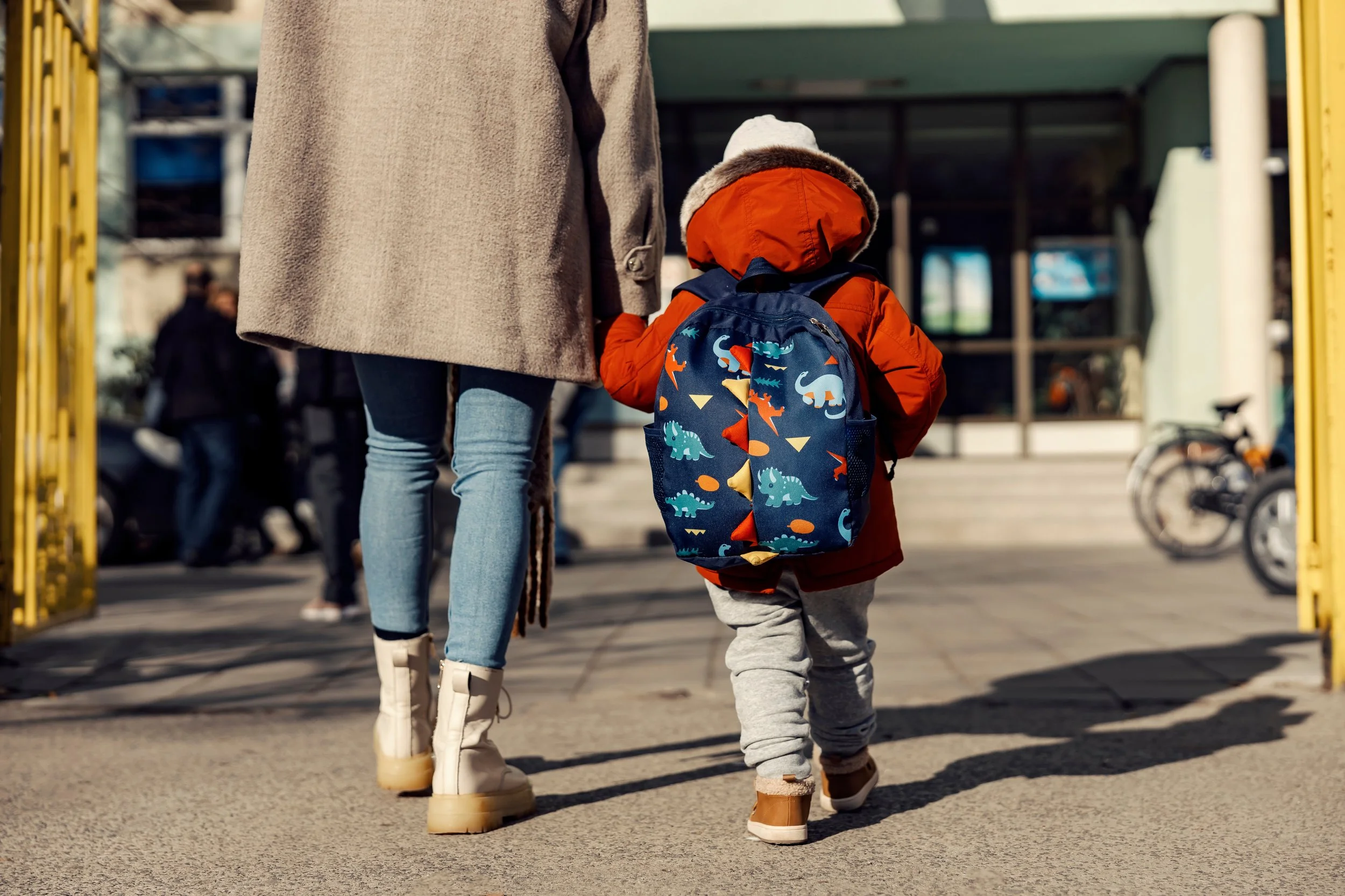 A woman and a young child walking through a public area, possibly a school or park, with the woman holding the child's hand. The child is wearing a red jacket, gray sweatpants, and a white hat, carrying a colorful dinosaur-themed backpack.
