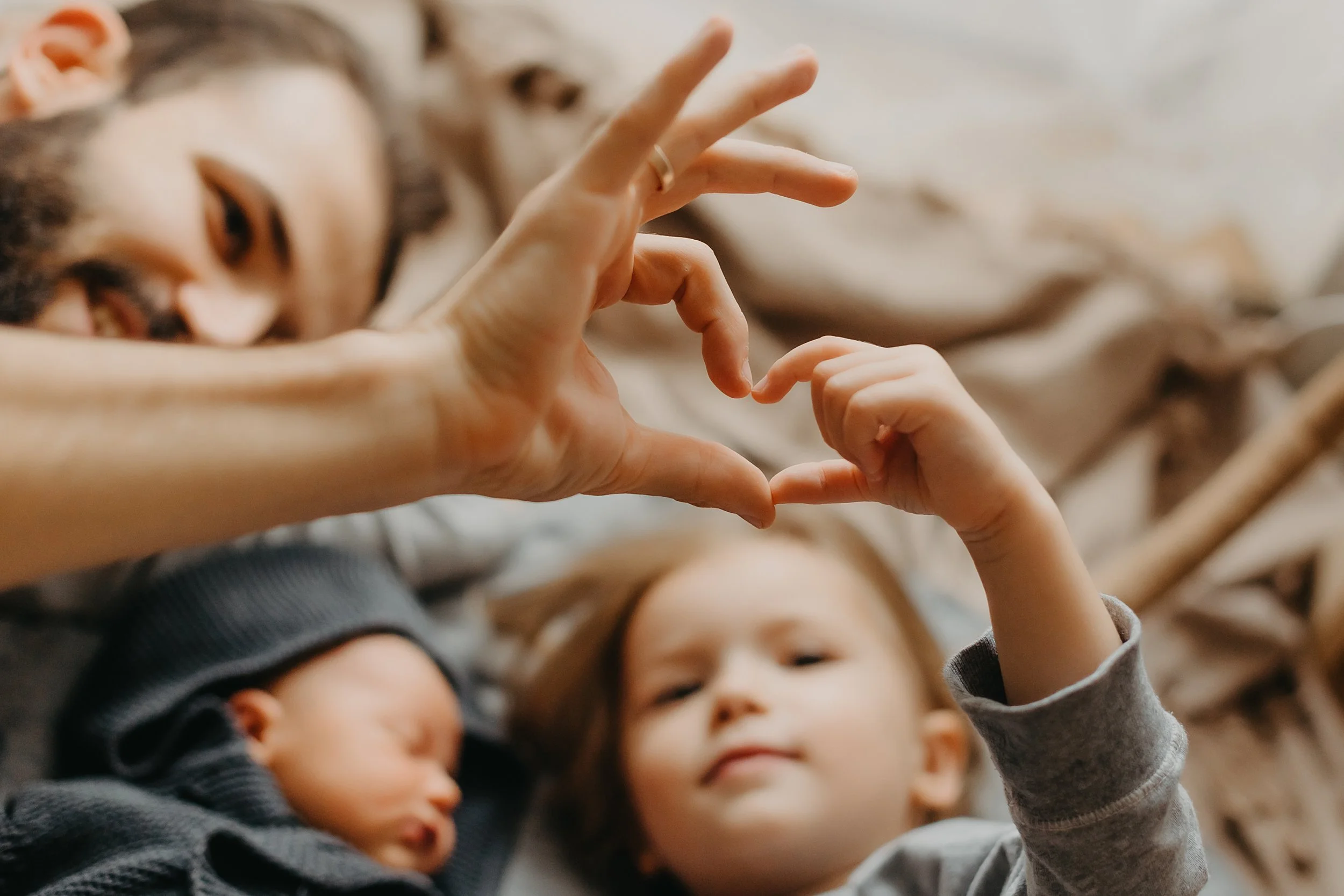 A man lying on a bed with two children, one sleeping and one awake, forming a heart shape with their hands.