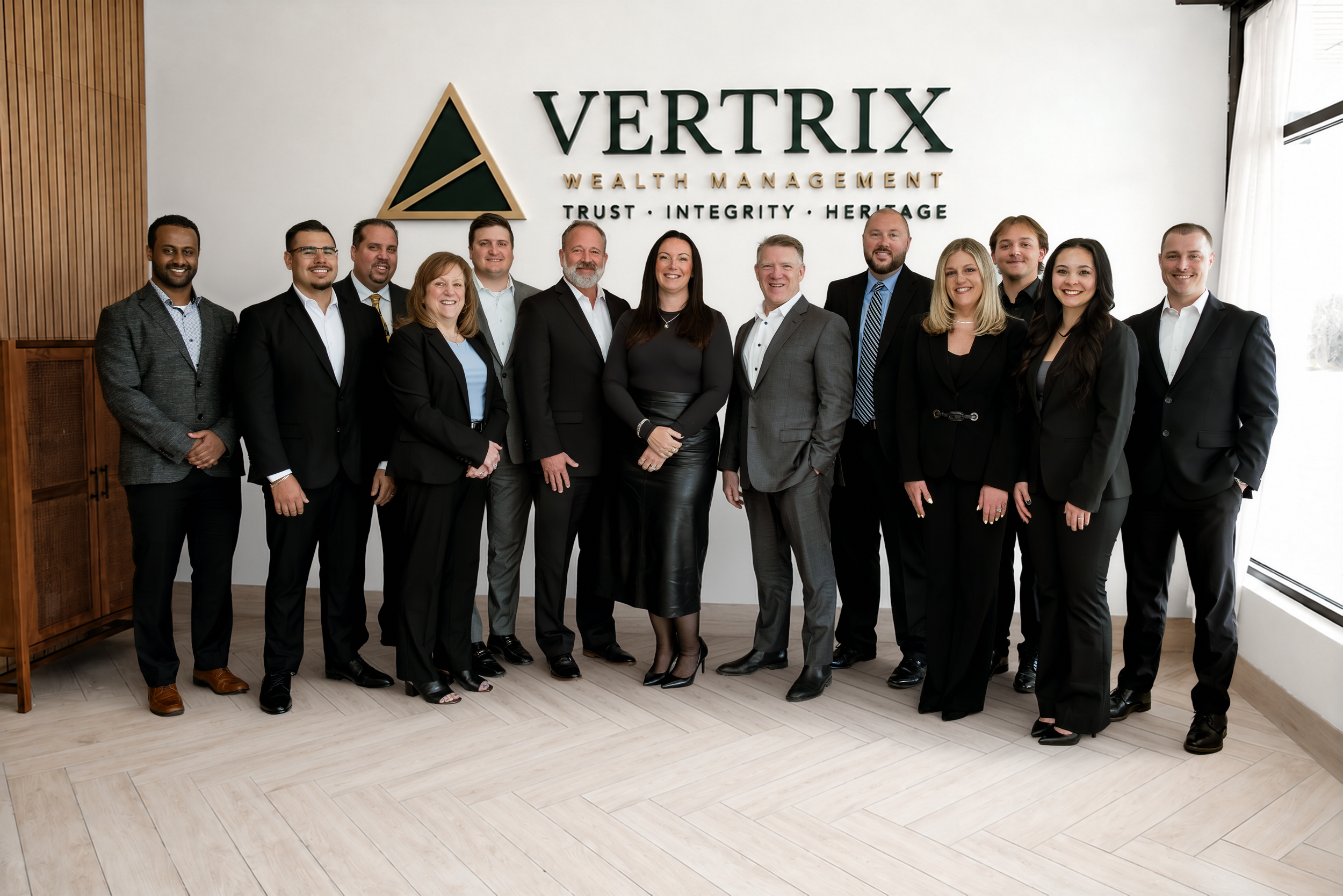 The Vertrix Wealth Management staff standing in front of the company logo sign on the wall of the office lobby.