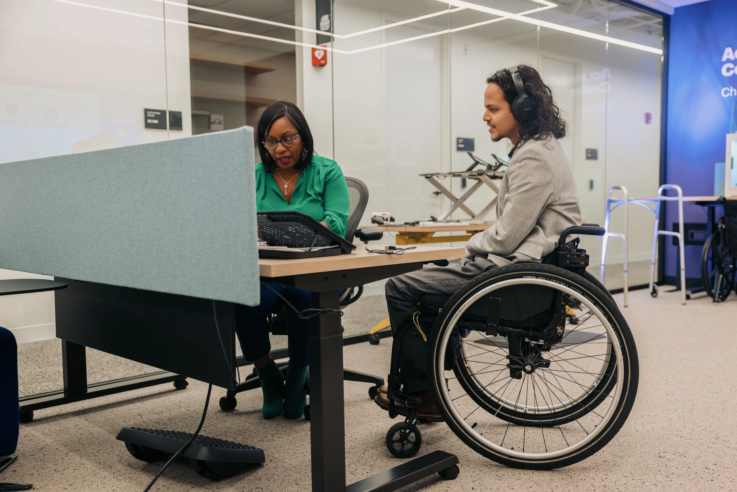 Two colleagues work together in an accessibility solutions center. One is wearing noise cancelling headphones and uses a wheelchair. The other reviews items with him on a laptop.