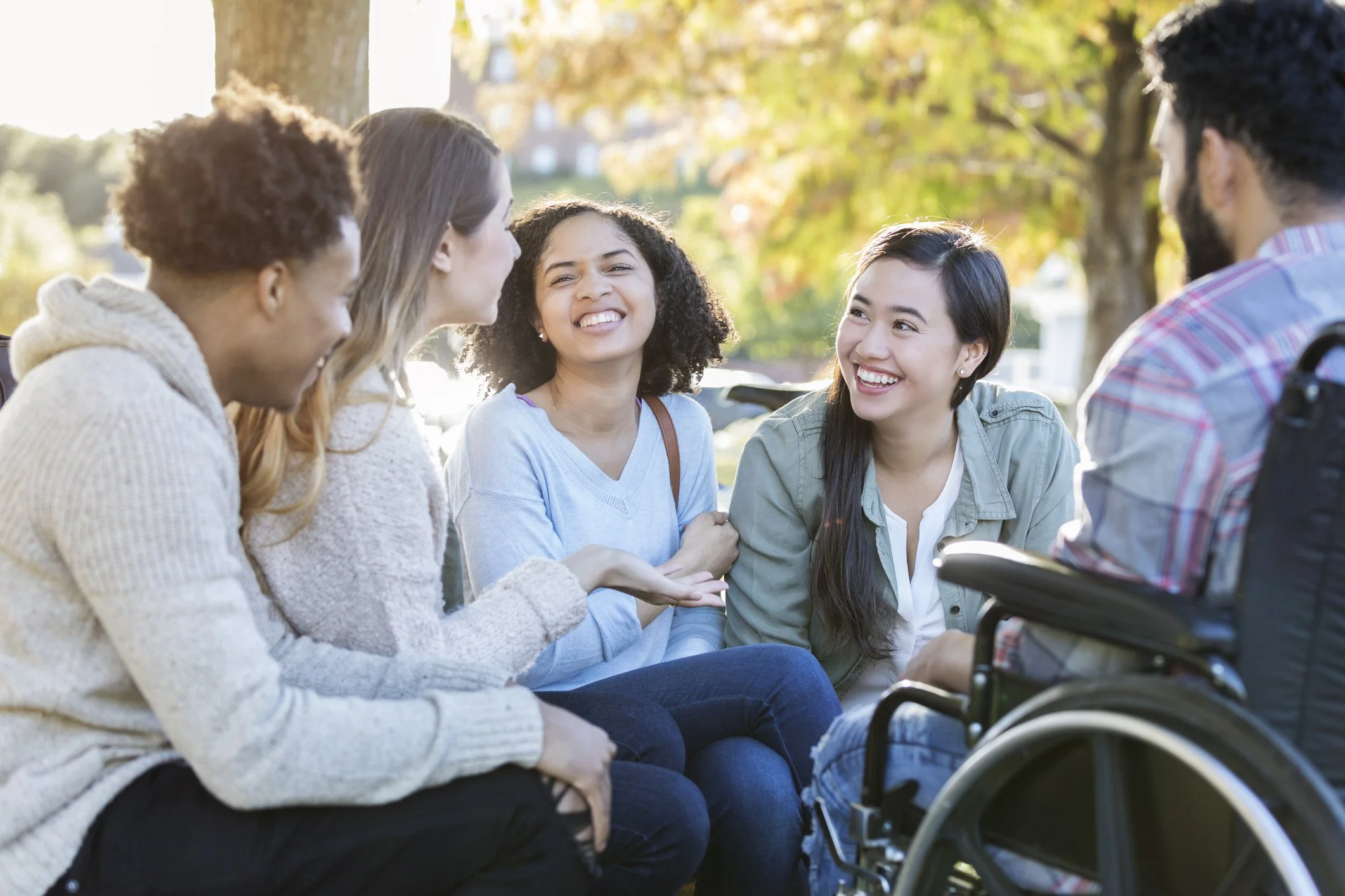 A group of college friends sit in a circle outdoors and enjoy a conversation. One member of the group, with his back to the camera, is a wheelchair user.