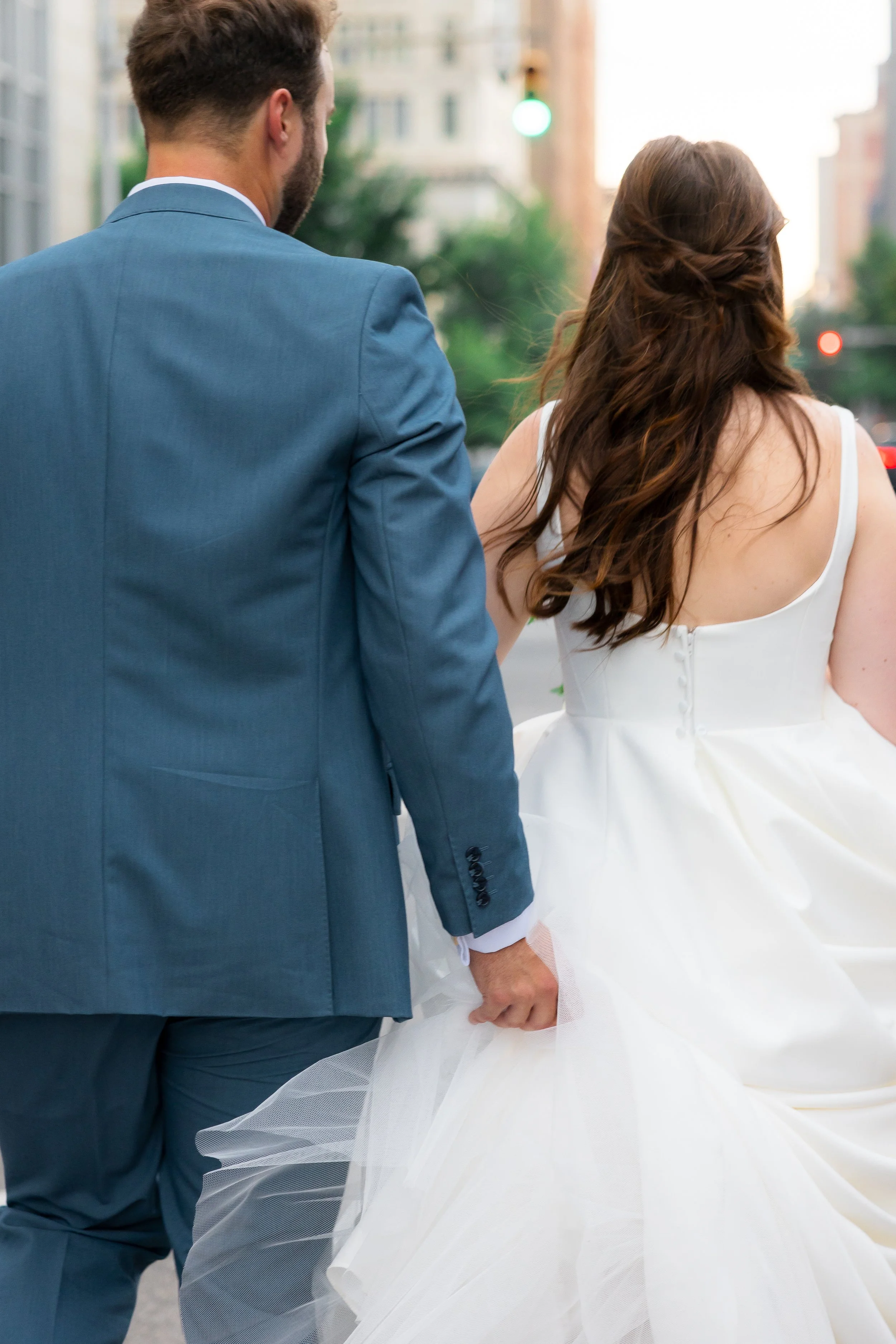 A bride and groom holding hands, walking together outdoors, with the bride in a white wedding dress and the groom in a blue suit.