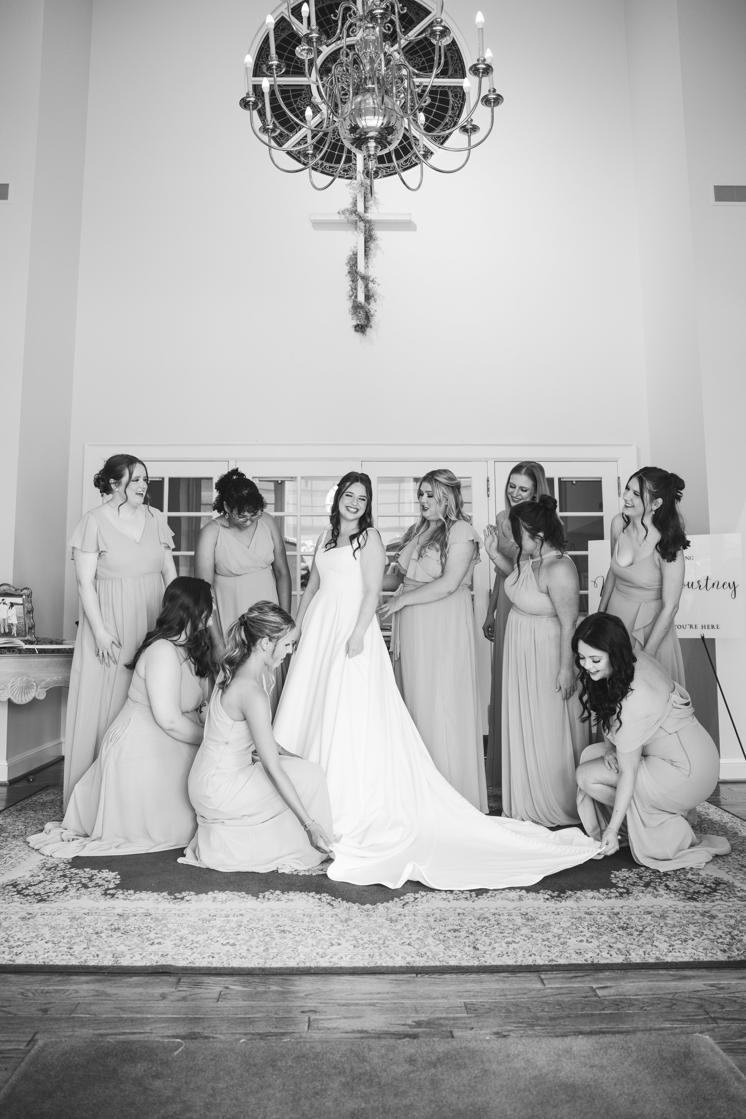 A bride with her bridesmaids preparing for her wedding, with some kneeling and others standing, in a room with a large chandelier on the ceiling.