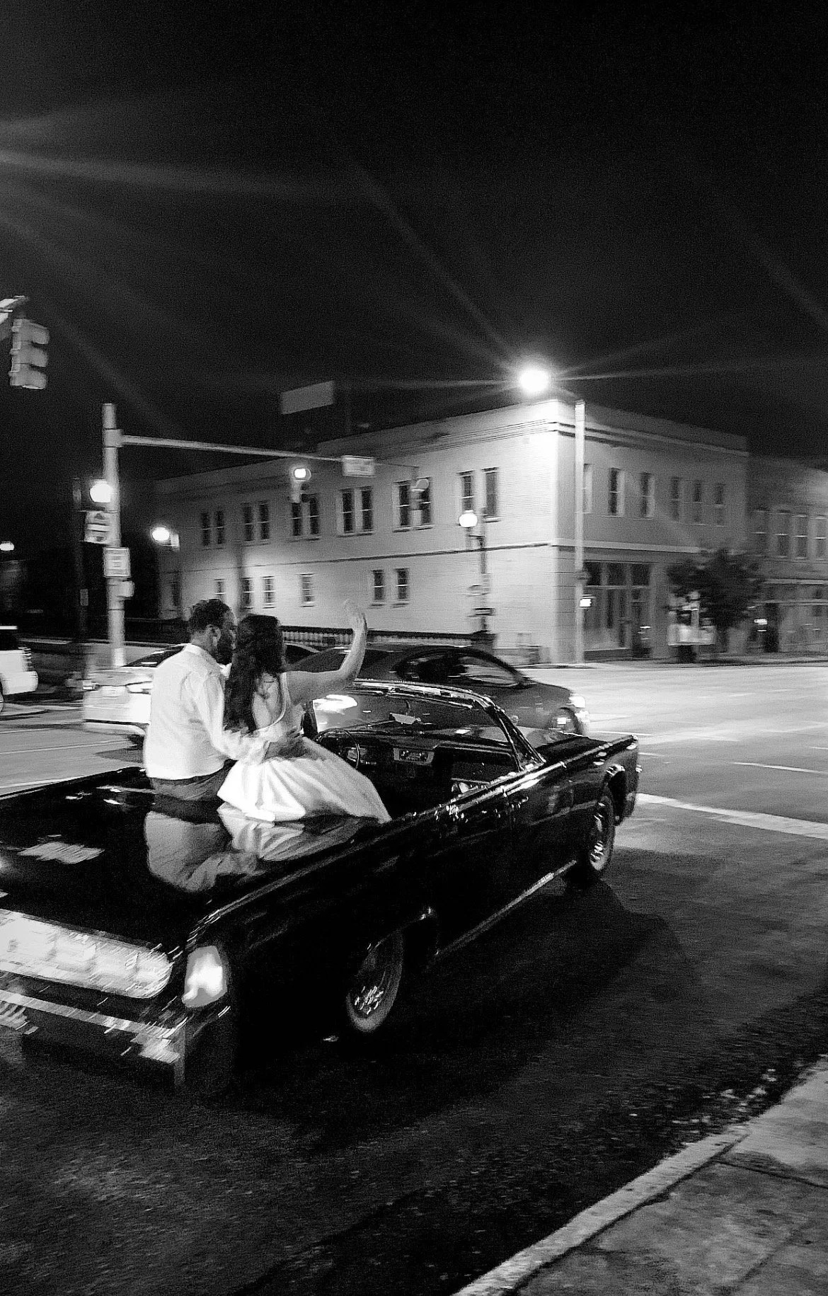 A black and white photo of a couple sitting in the back of a vintage car at night, with one person waving, on a city street with buildings, streetlights, and passing cars in the background.
