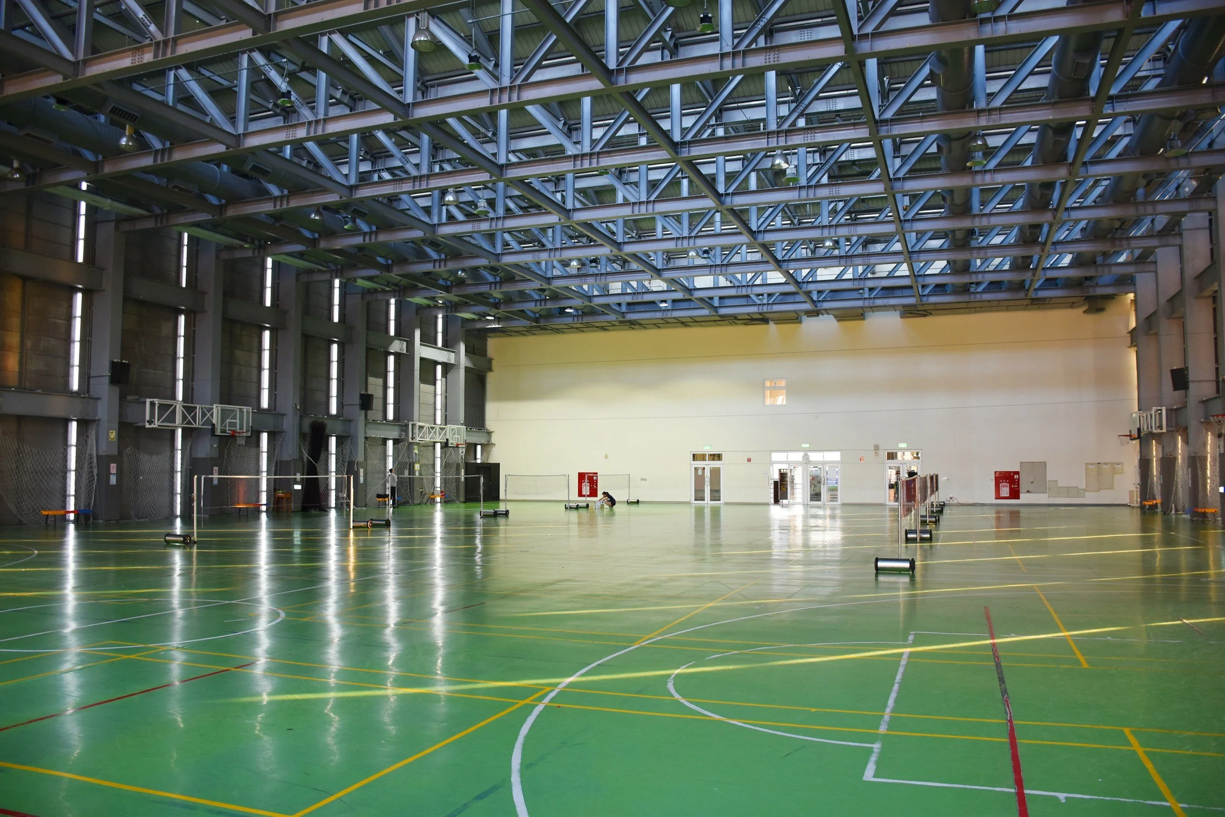 An indoor gymnasium with a green sports floor, several volleyball nets, and basketball hoops. The ceiling has a steel truss structure with industrial lighting. The back wall has doors and safety equipment.