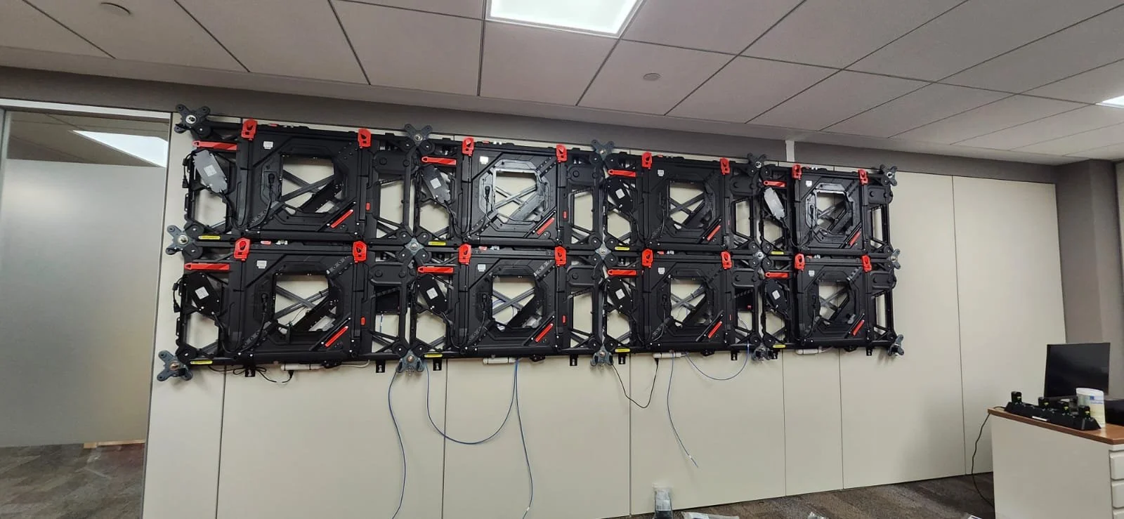 A large wall-mounted LED display panel with a black and red framework, connected with wires, in an office room.