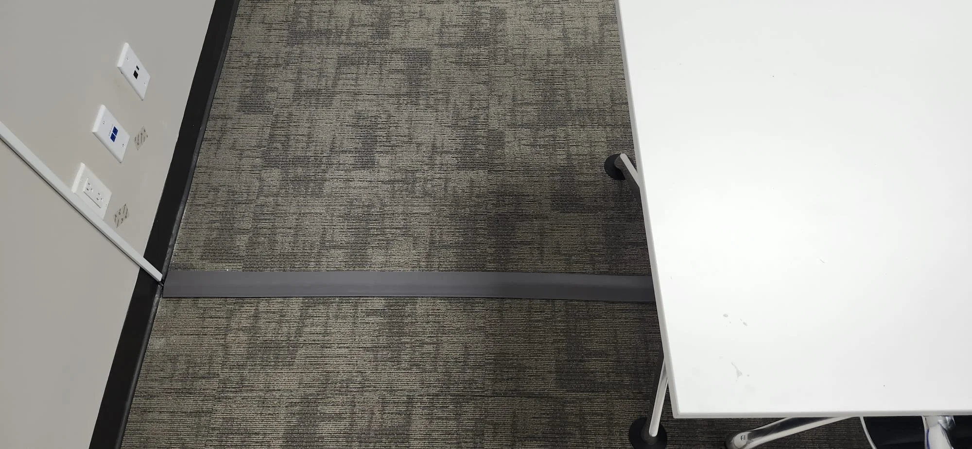 Part of an office or conference room showing a white table, a textured carpeted floor, and electrical outlets and network ports on a beige wall.