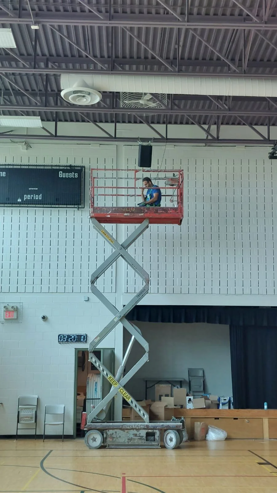 A person standing on a red scissor lift inside a gymnasium, holding a phone or device. The gym has a high ceiling with visible ductwork and a scoreboard on the wall.
