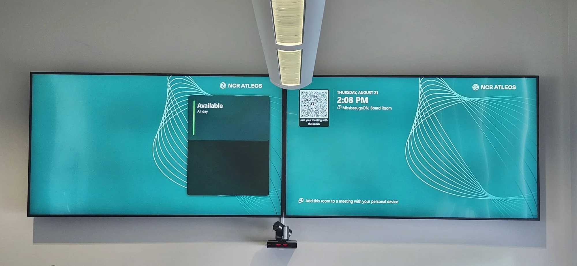 Digital display screen showing meeting room availability, time, date, and QR code for joining a virtual meeting at a corporate office.