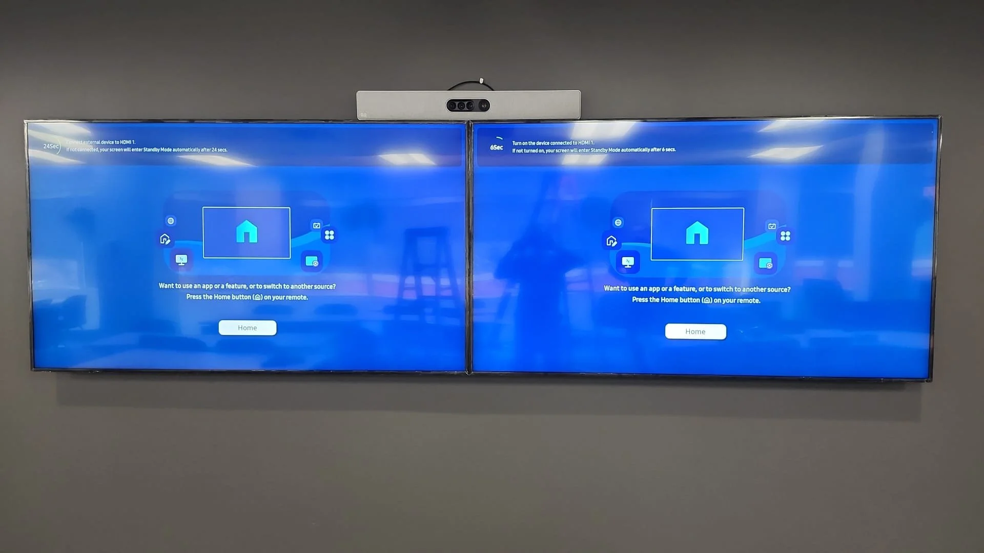 Two large flat-screen monitors mounted on a gray wall displaying a blue setup screen with a home icon in the center, instructions, and a 'Home' button.