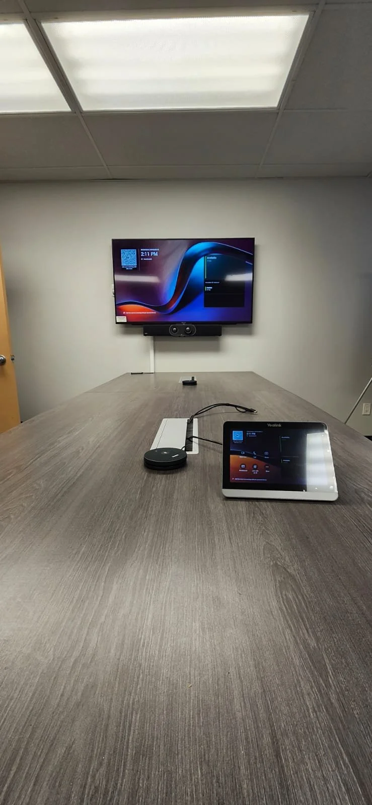 Conference room with a wooden table, a large wall-mounted flat screen TV, and a tablet device connected to a wireless speaker on the table.