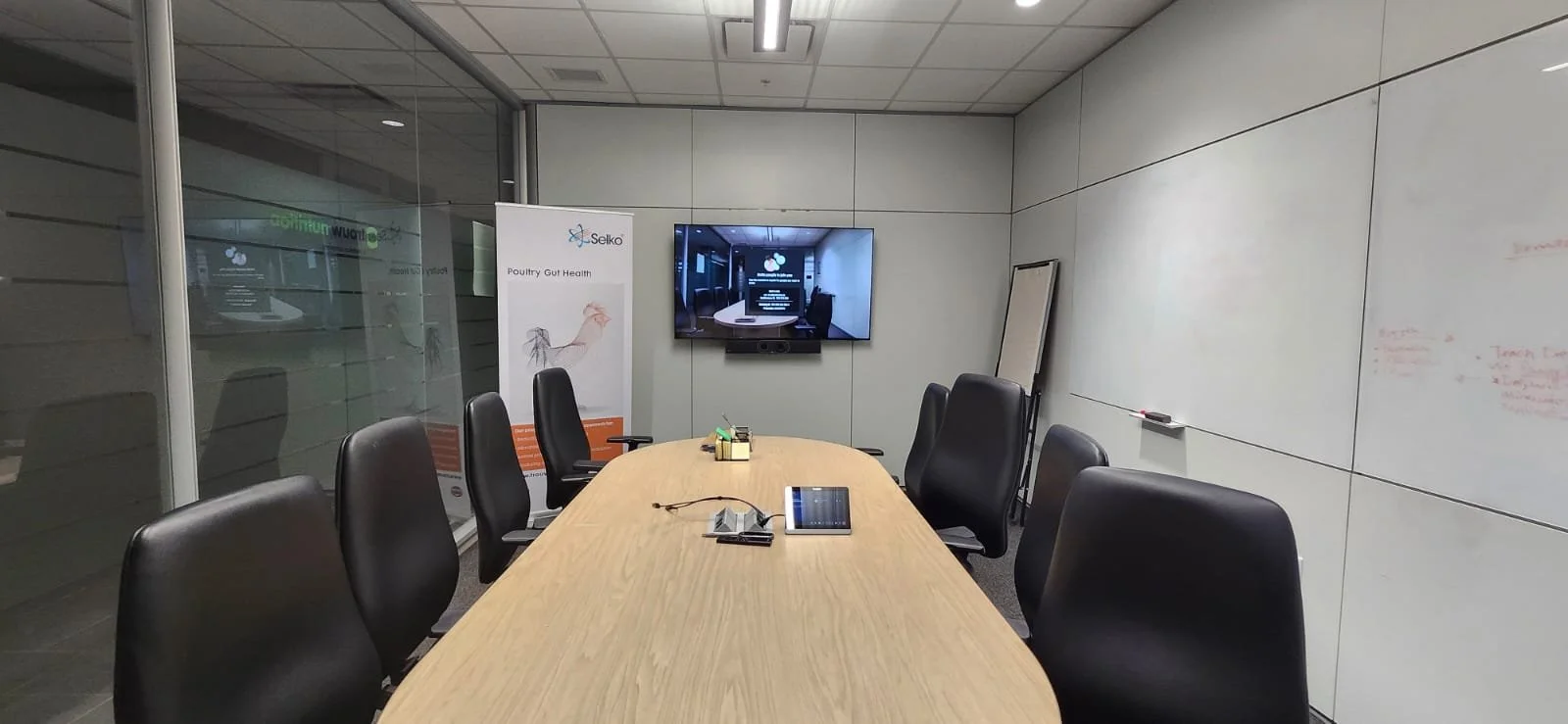 Empty conference room with a long oval wooden table, eight black office chairs, a TV screen on the wall, a whiteboard, and a promotional stand for poultry gut health.
