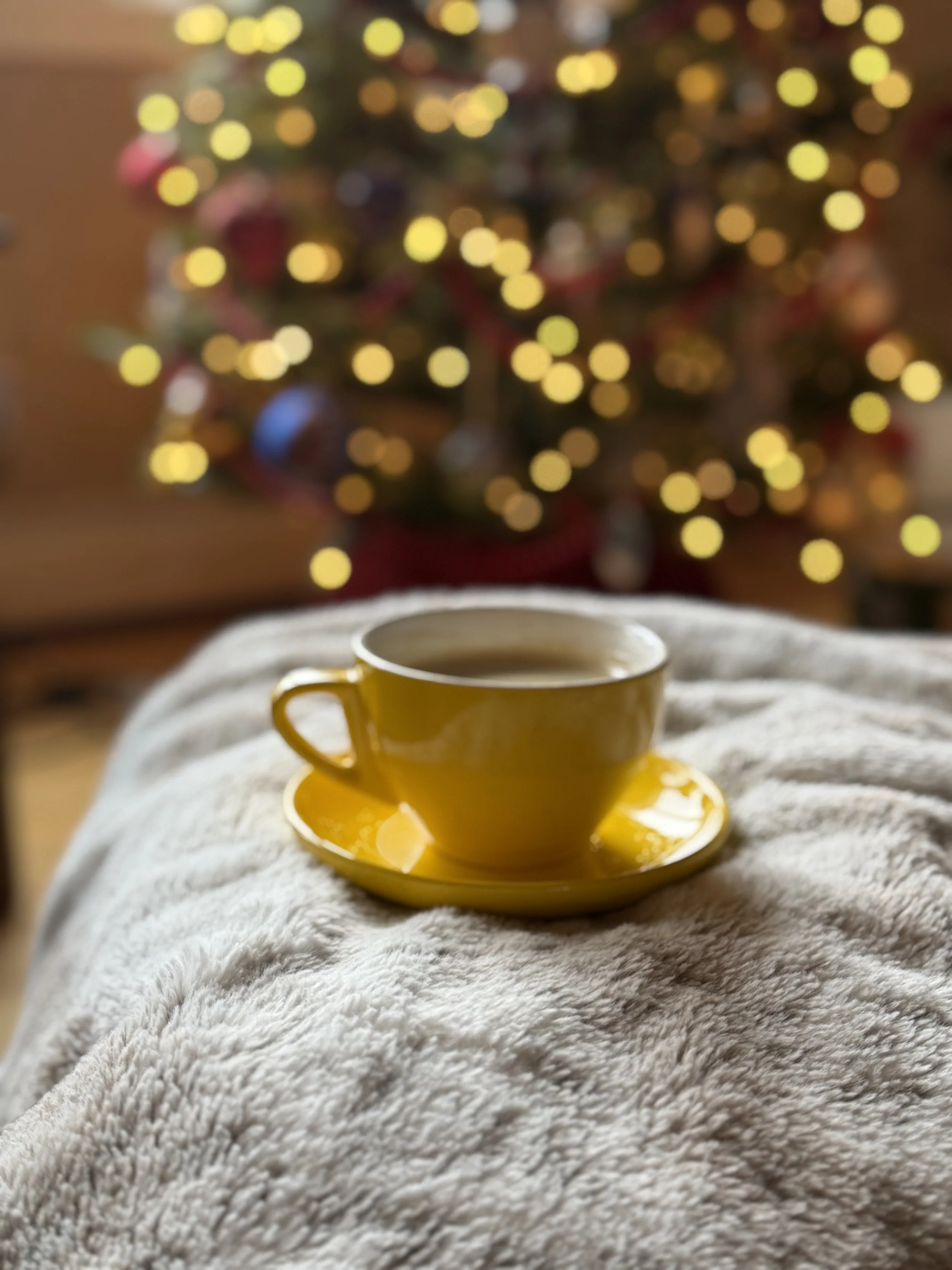 A yellow cup and saucer on a cozy, white, plush blanket with Christmas tree blurred in the background