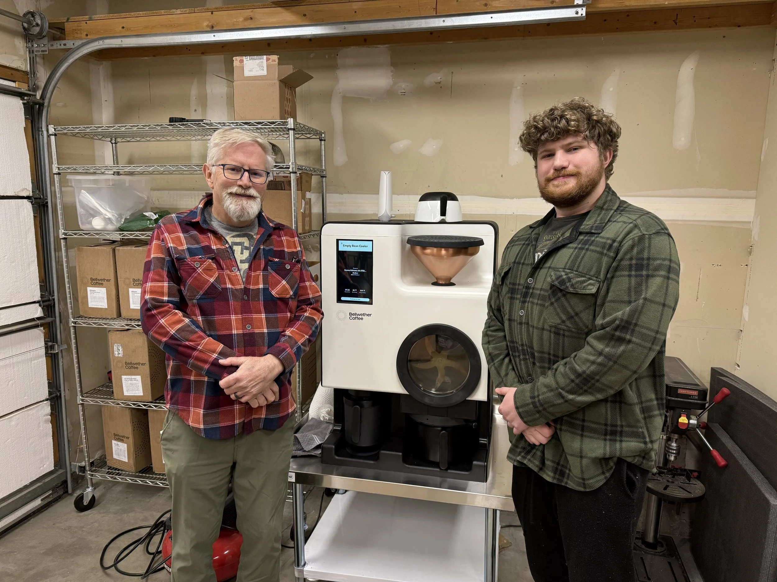 Two men standing in a garage near a coffee roasting machine, with shelves of boxes in the background.