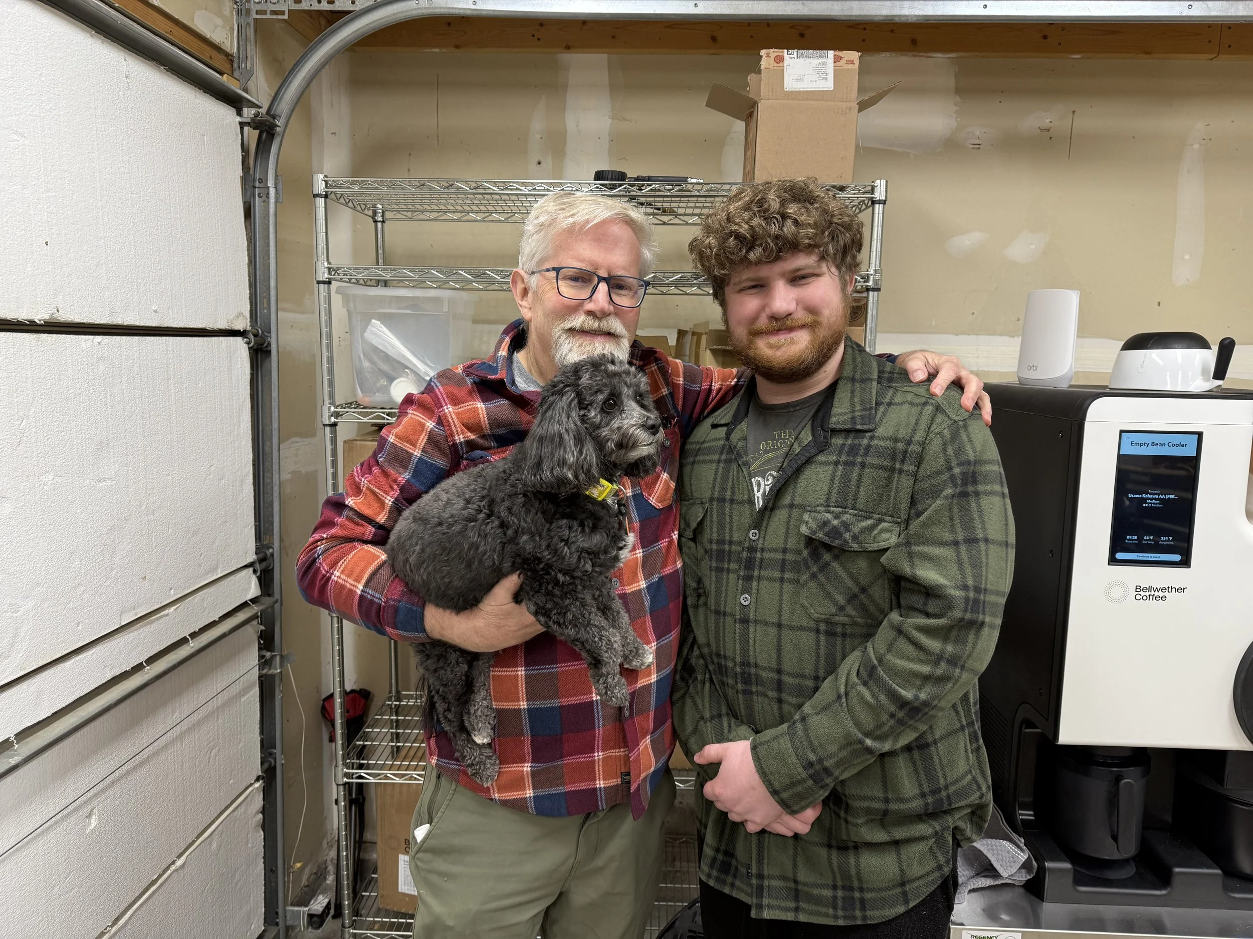 Two men, one older with glasses and a beard, and one younger with curly hair and a beard, standing in a garage with a small black dog. The older man is holding the dog, and both are smiling. A metal shelf, a box, and a coffee machine are visible in the background.
