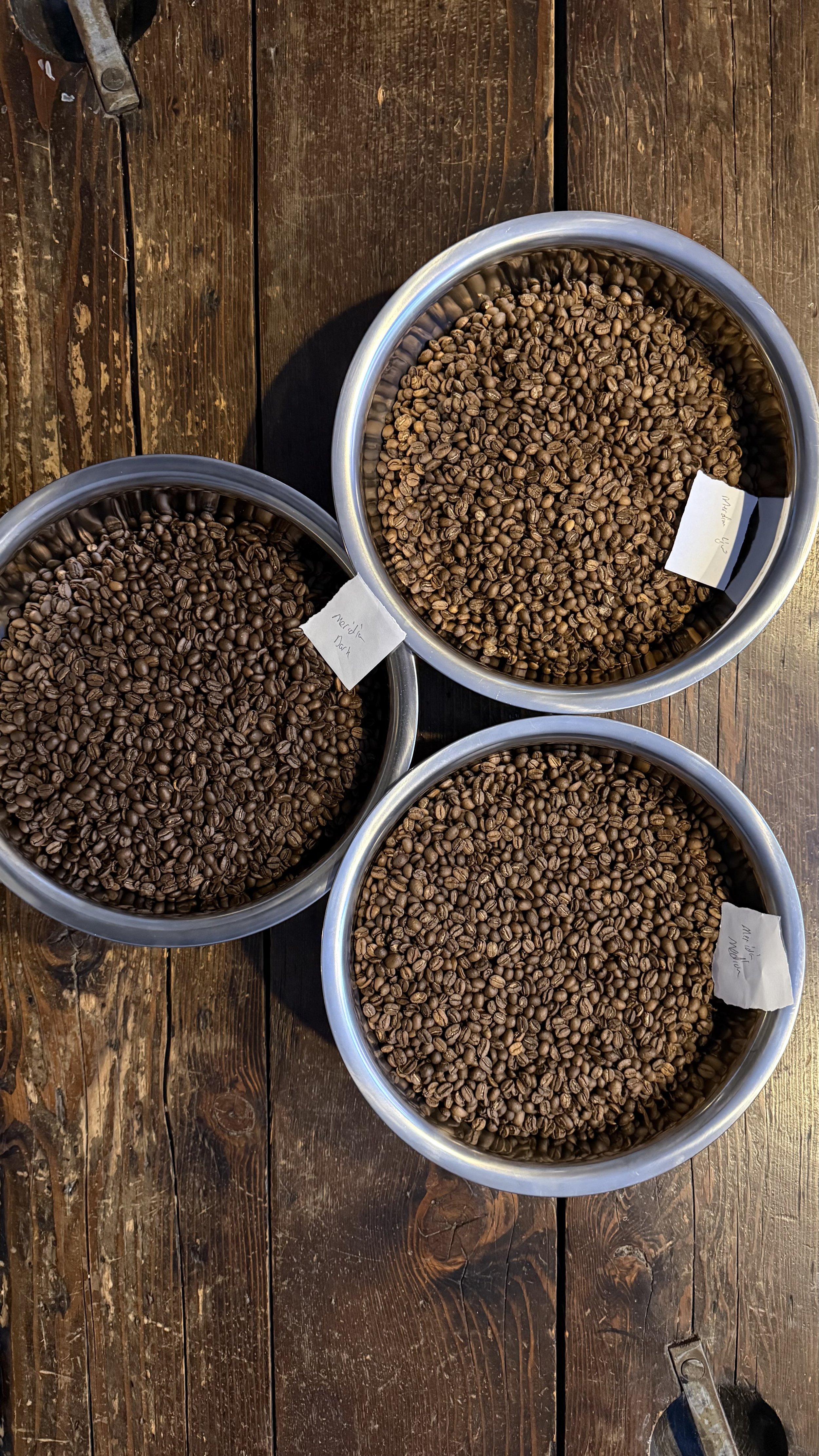 Three metal bowls filled with different types of roasted coffee beans placed on a wooden surface.