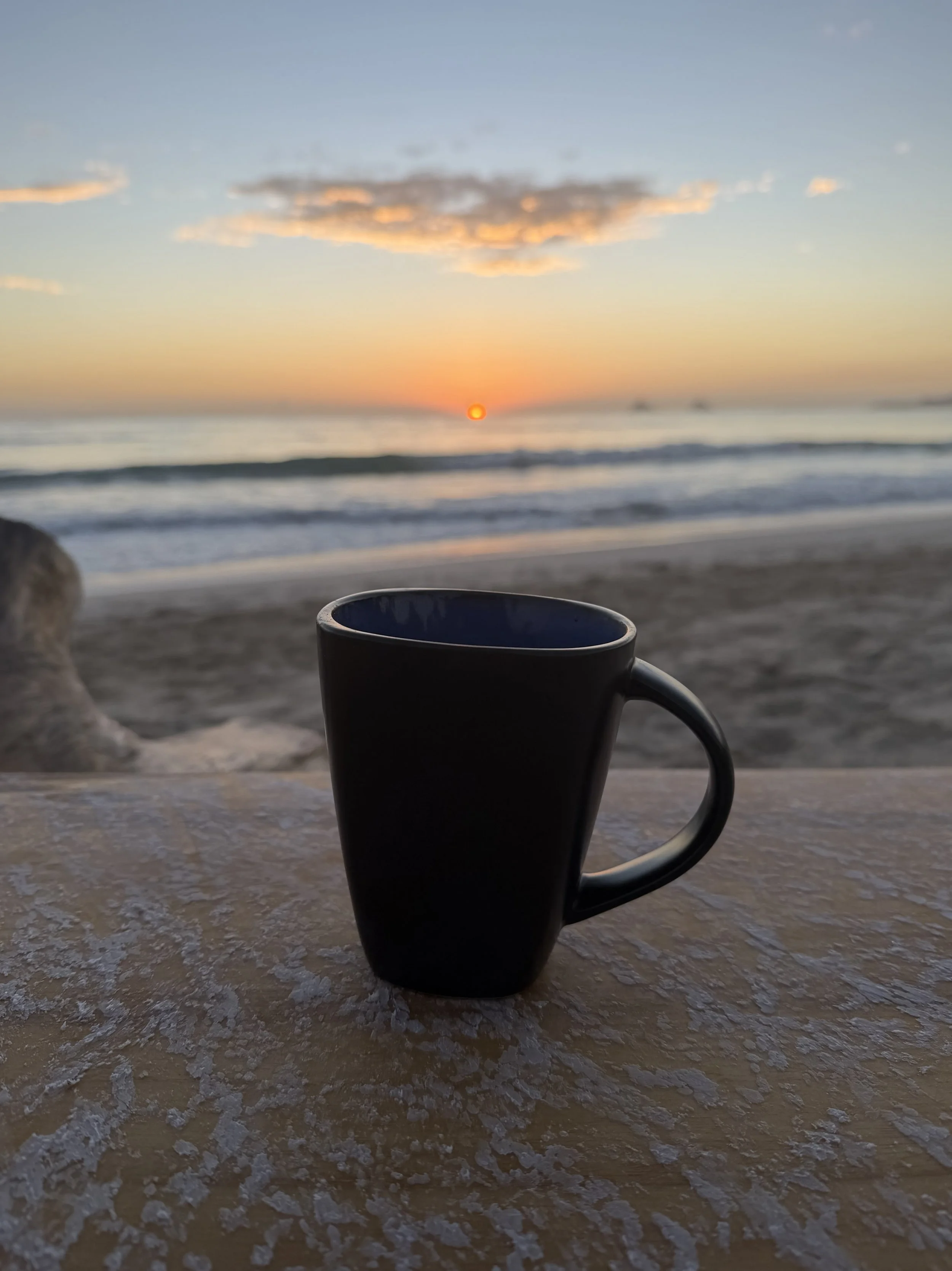 Black coffee mug on a textured surface with a scenic beach sunset in the background, ocean waves and a partly cloudy sky.