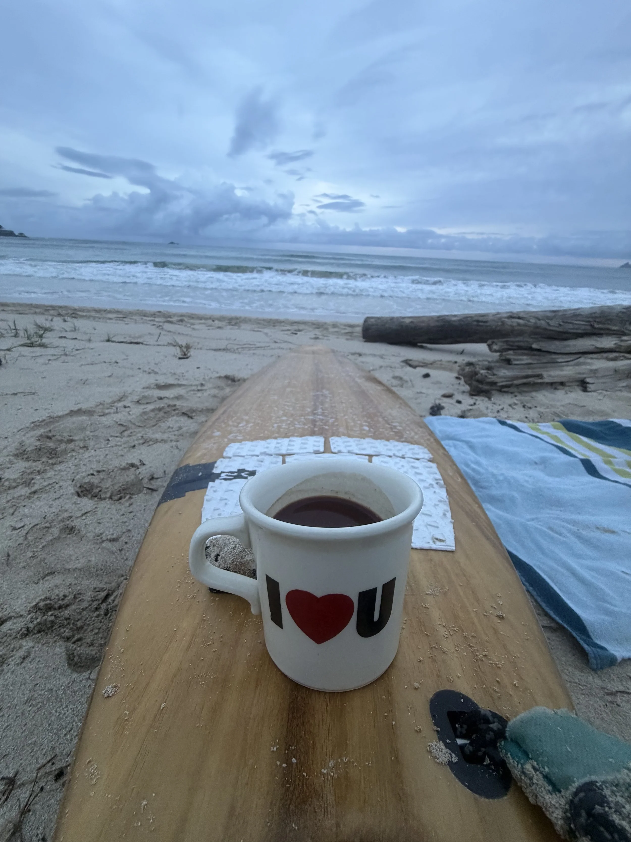A cup of coffee on a paddleboard at the beach, with the ocean, waves, driftwood, and cloudy sky in the background.