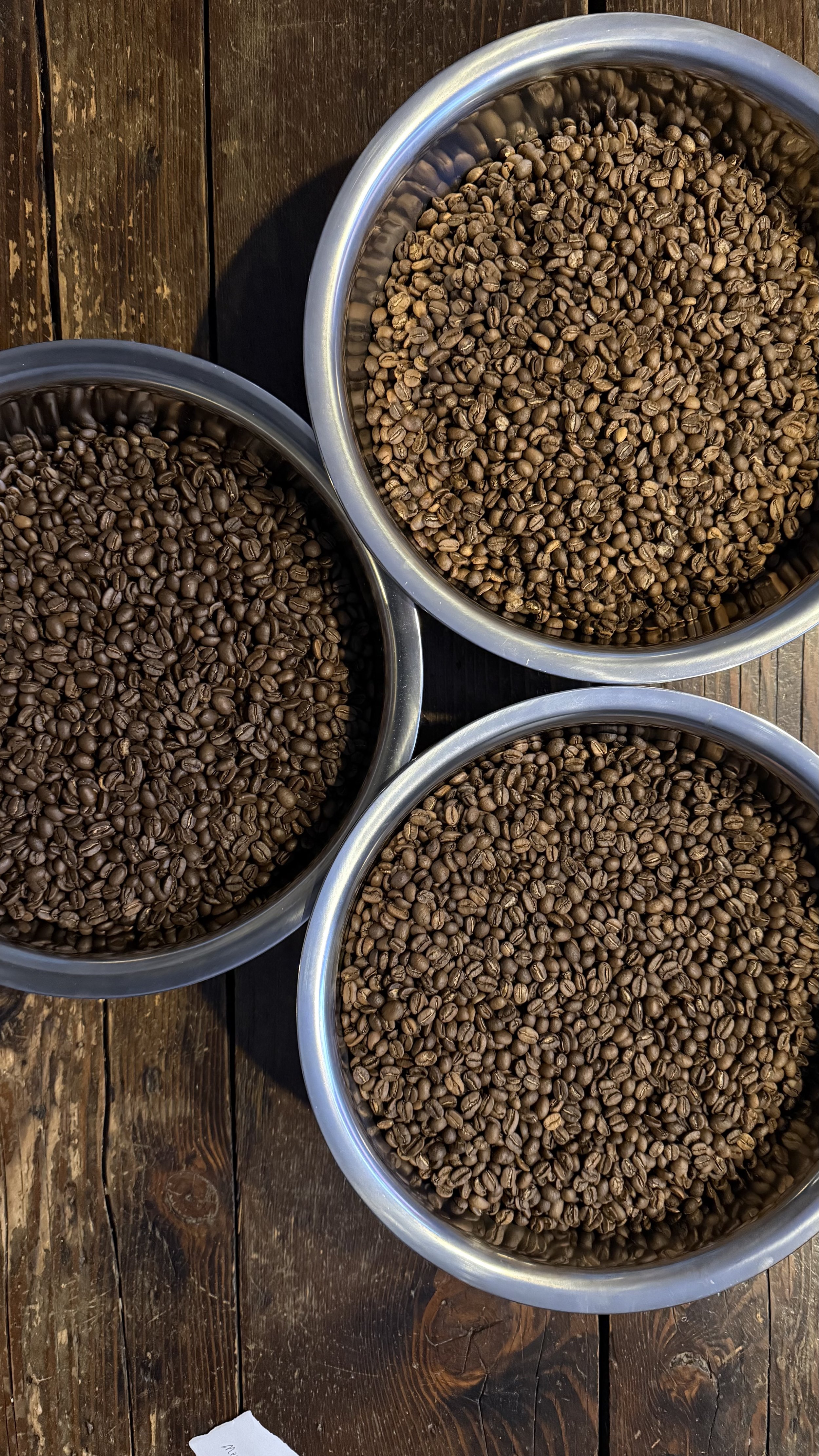Three metal bowls filled with different types of coffee beans on a wooden surface.
