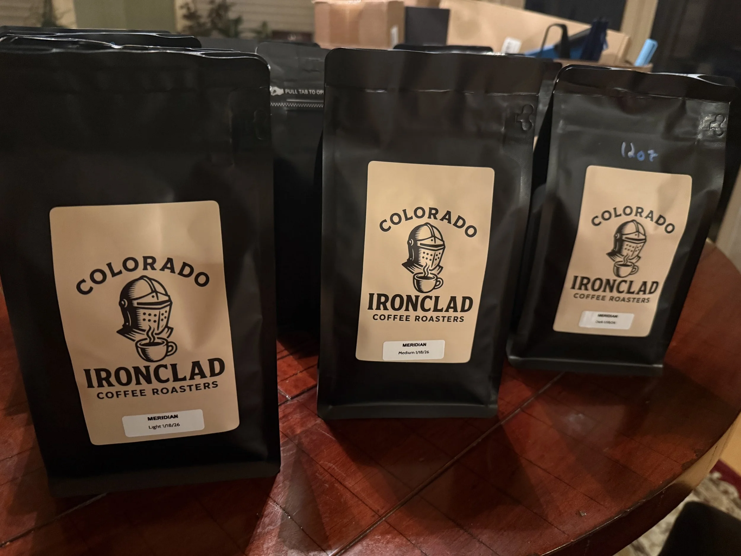 Three black coffee bags labeled 'Colorado Ironclad Coffee Roasters' on a wooden table. The bags feature a logo of a knight's helmet with a steaming coffee cup below. The left bag is labeled 'Meridian, Light 1/8/26', the middle 'Meridian, Medium 1/8/26', and the right one 'Meridian, Dark 1/8/26'.