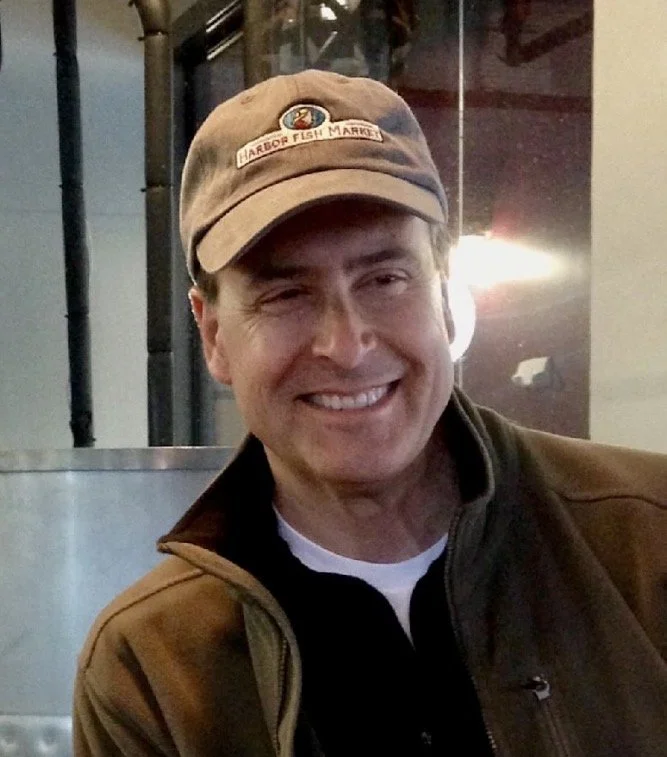 A smiling man wearing a baseball cap and brown jacket indoors.