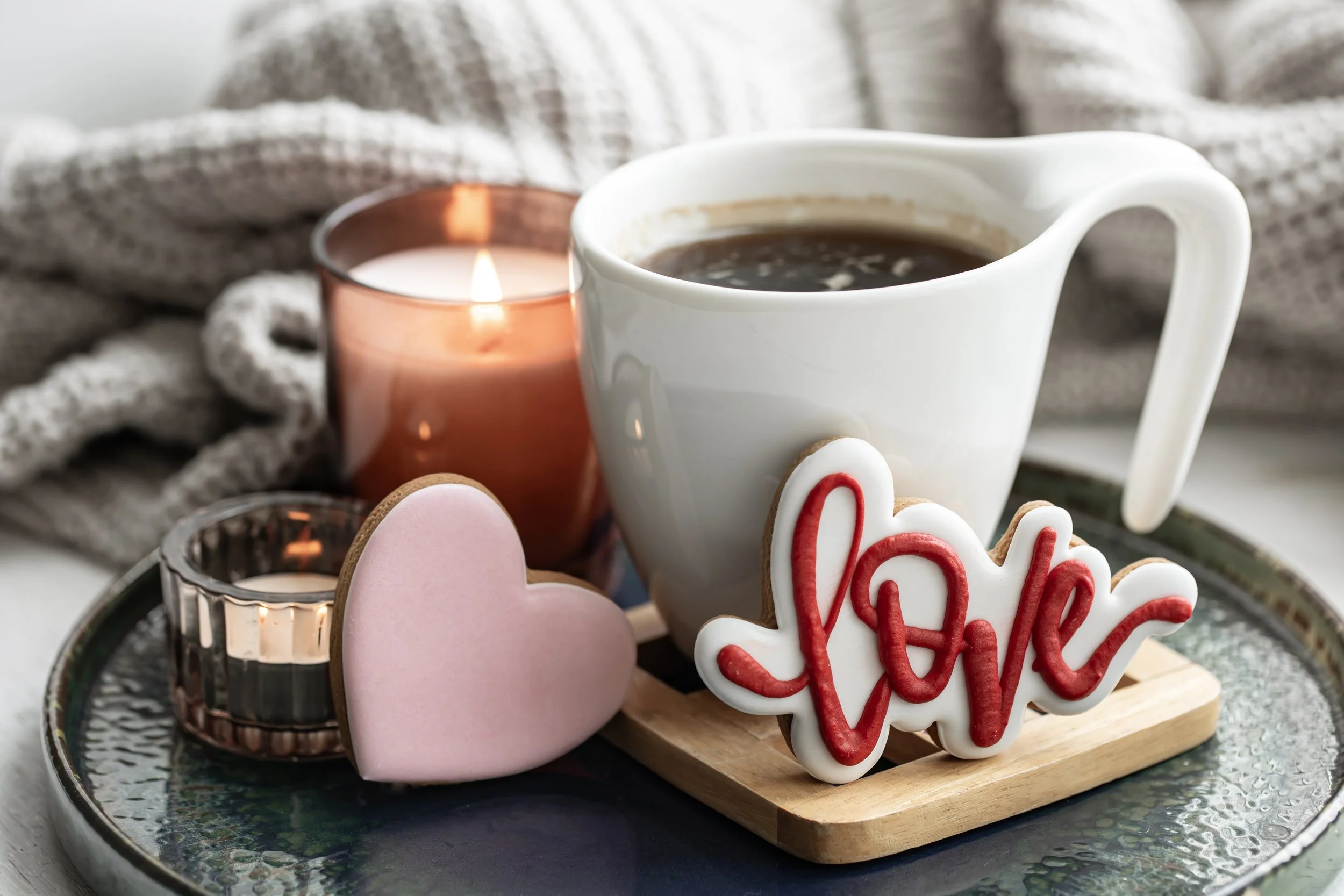 A tray with a white mug of coffee, a pink heart-shaped cookie, a white 'love' cookie with red icing, a small candle, and a copper candle holder, with a cozy gray blanket in the background.