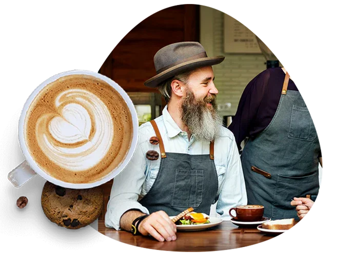 A barista with a beard and hat smiles at a customer while sitting at a coffee shop counter, with a latte featuring latte art, cookies, and a cup of coffee on the table.