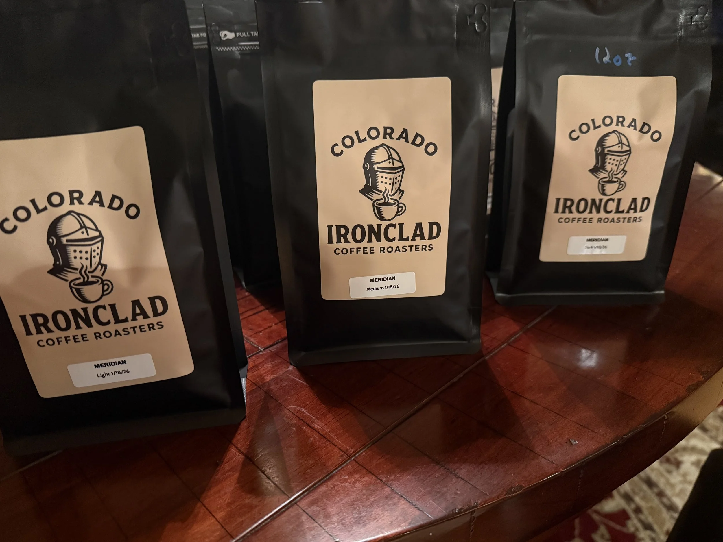 Three bags of Colorado Ironclad Coffee Roasters on a wooden table, labeled as Meridian Light, Meridian Medium, and Meridian Dark roast.