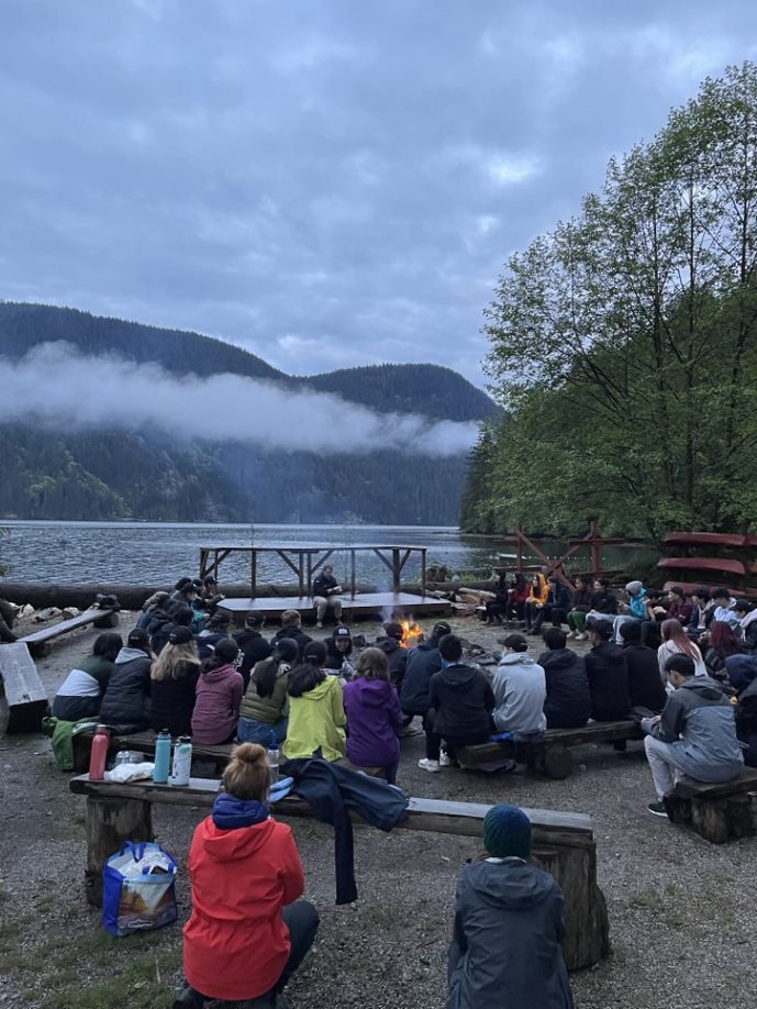 A group of people sitting around a campfire on a beach near a lake, with hilly and forested landscape in the background.