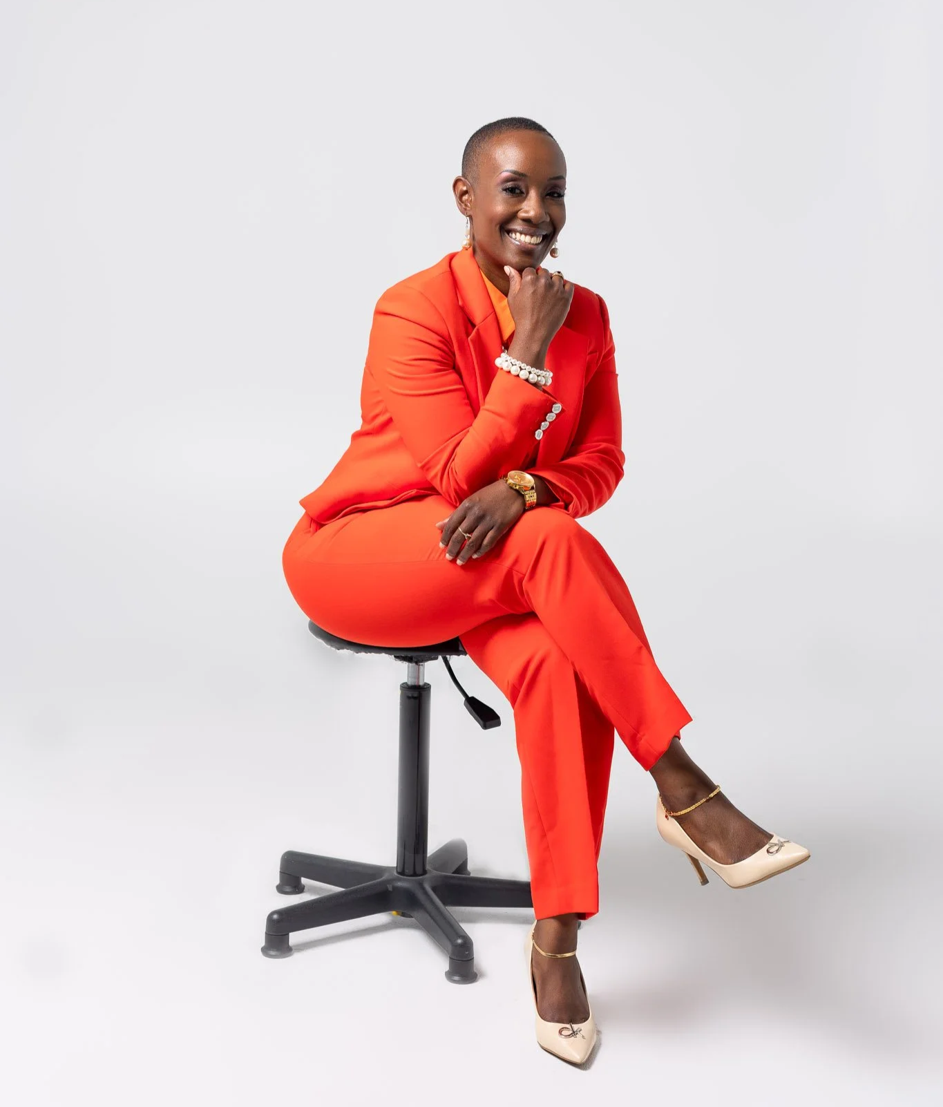A woman in an orange suit sitting on a black office stool, smiling, with her right hand resting on her chin, against a plain white background. She is accessorized with pearl jewelry and beige high heels.