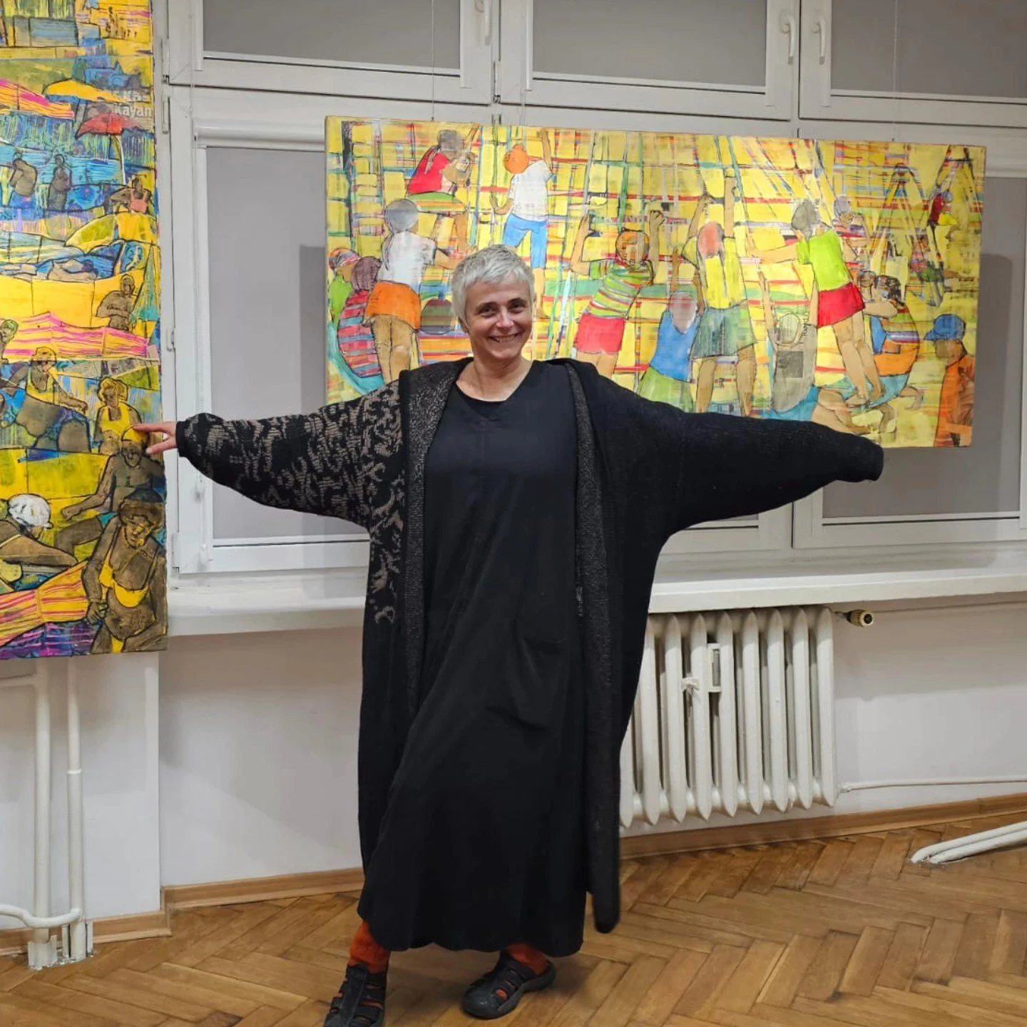A smiling woman with short gray hair wearing a long black dress and patterned black cardigan, standing in front of colorful paintings featuring scenes of people at a beach and a library.