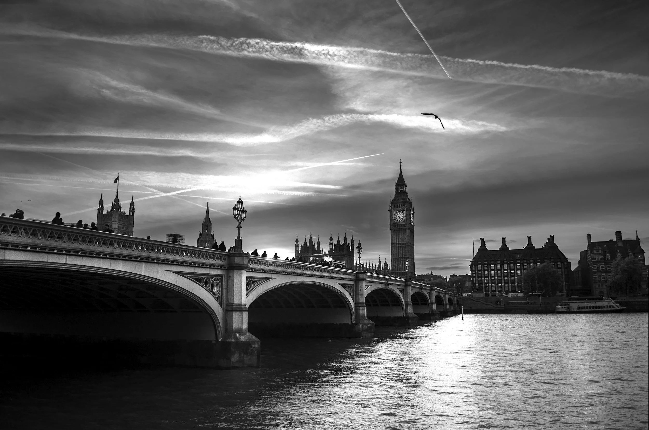 Black and white photo of Westminster Bridge spanning the River Thames in London, with the Big Ben clock tower in the background. There are a few birds flying in the sky and some people on the bridge.