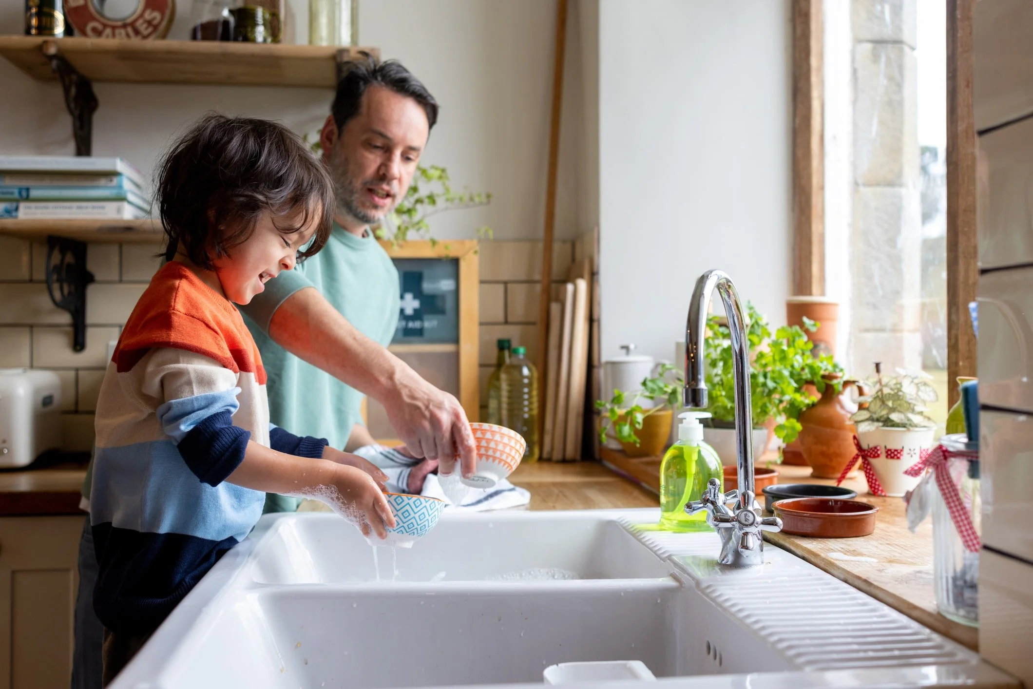 A boy and a father doing dishes at a kitchen sink, with the boy smiling and the man watching, in a bright, cozy kitchen with plants and sunlight.