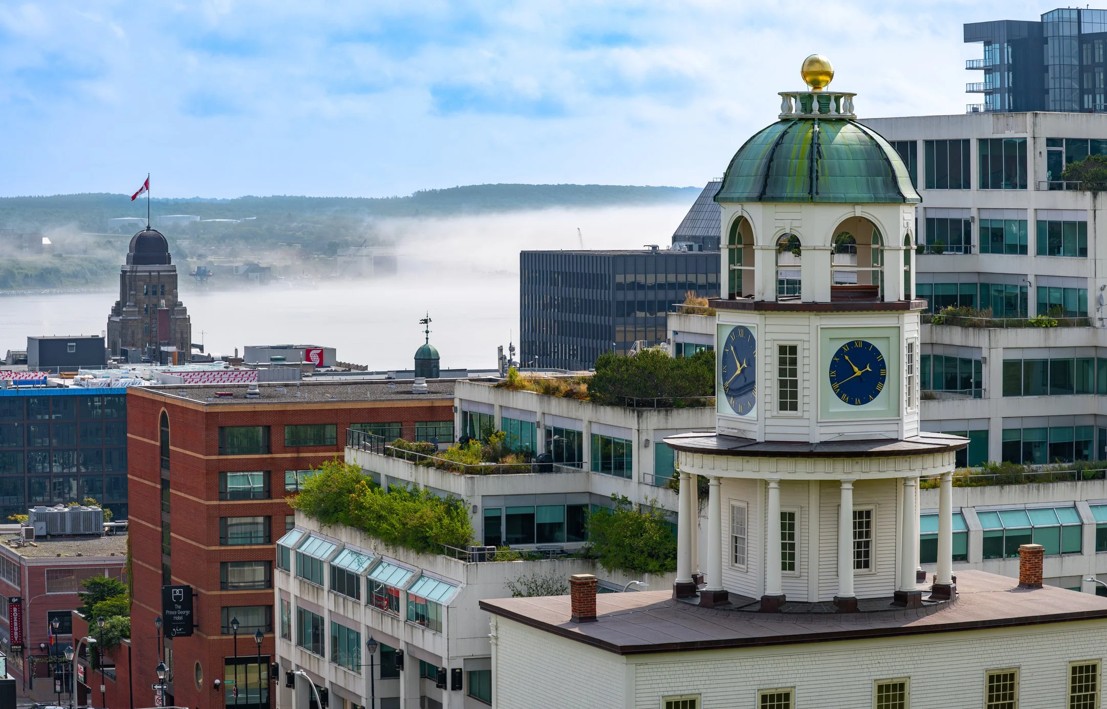Cityscape with a historic clock tower in the foreground, modern buildings, and a body of water with mist in the background.