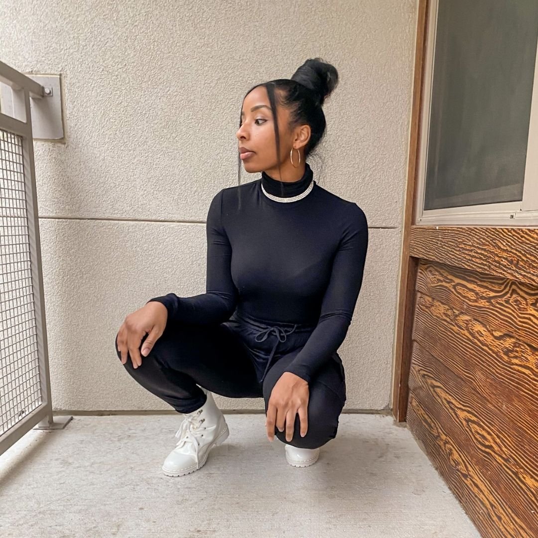LaNese on balcony with black turtle neck, joggers, white boots, and statement necklace