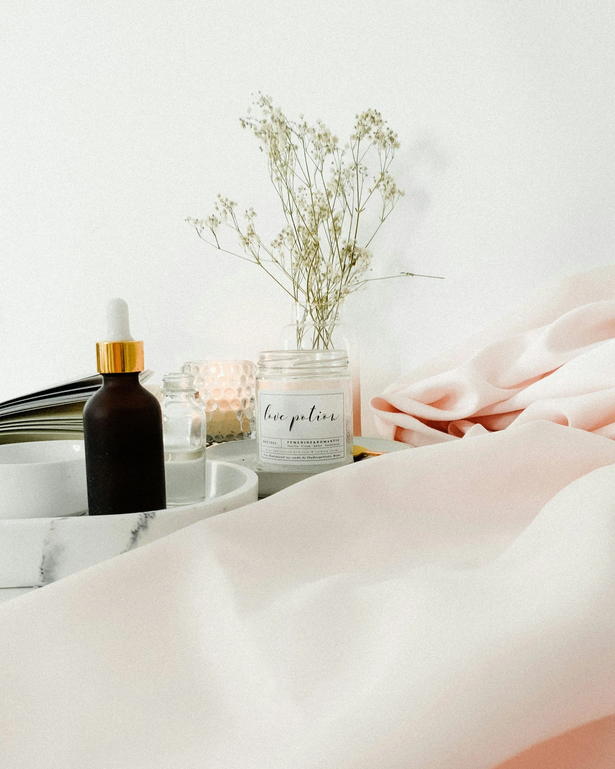 A white surface with a black bottle with a gold cap, a book, a pink candle, and a glass jar with white flowers, with soft pink fabric on the right side.